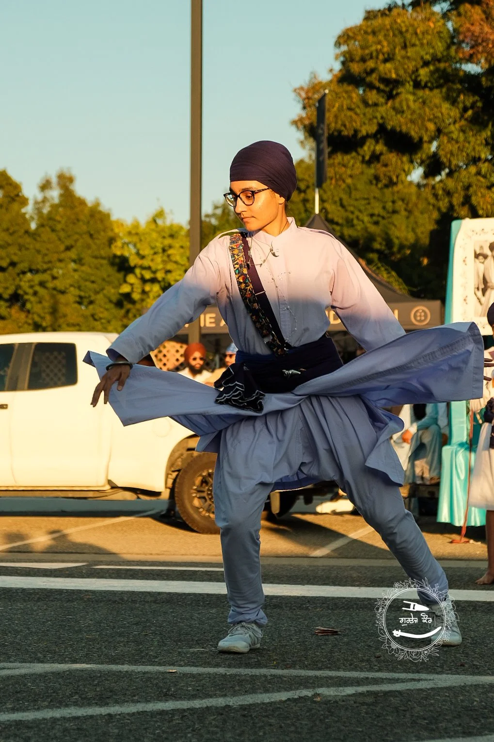 A woman dressed in traditional Sikh attire, including a black turban, black glasses, and a white kurta, performing a dance or martial arts move on a city street during daytime.