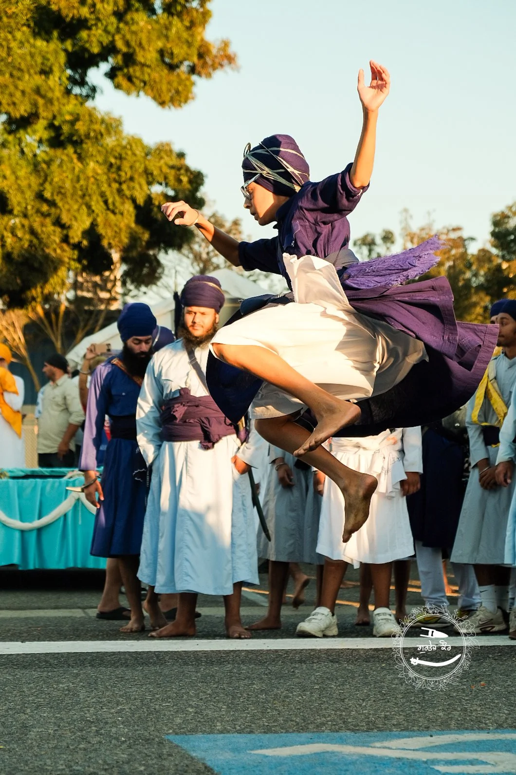 A man in traditional Sikh attire jumps in the air during a cultural celebration, with other similarly dressed men and a float in the background.