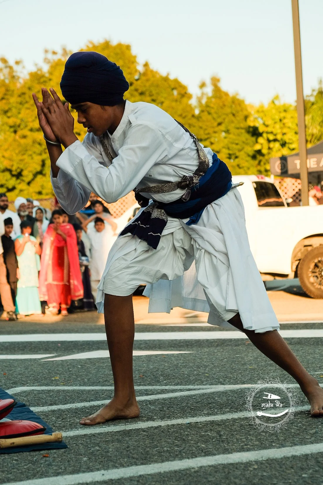 A young man dressed in traditional white attire with a blue sash and head covering practices martial arts or a cultural ritual on a street, with spectators in the background.