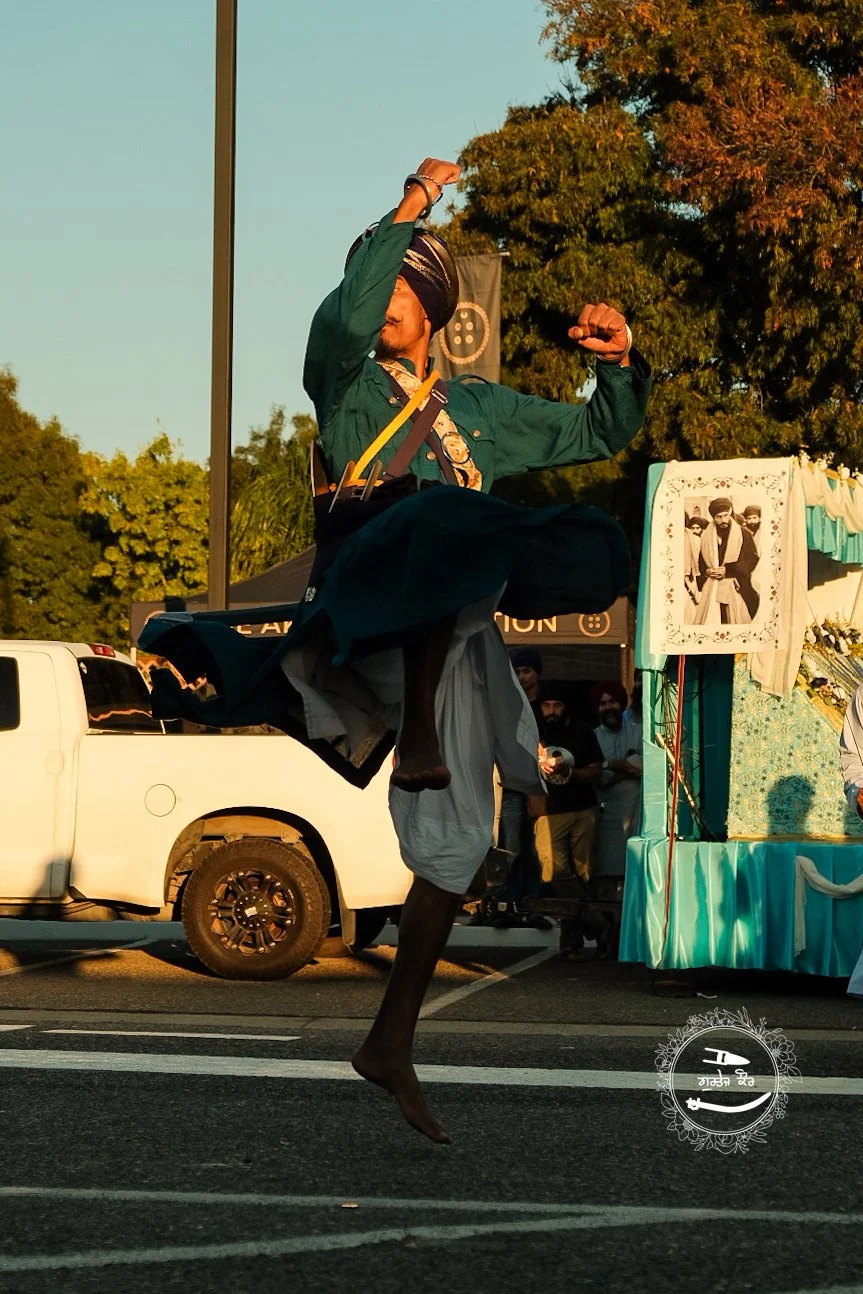 A person dressed in traditional Sikh attire performing a jump in the air during a parade or celebration, with a float decorated with a portrait of Sikh figures in the background, and other participants and a white pickup truck visible.