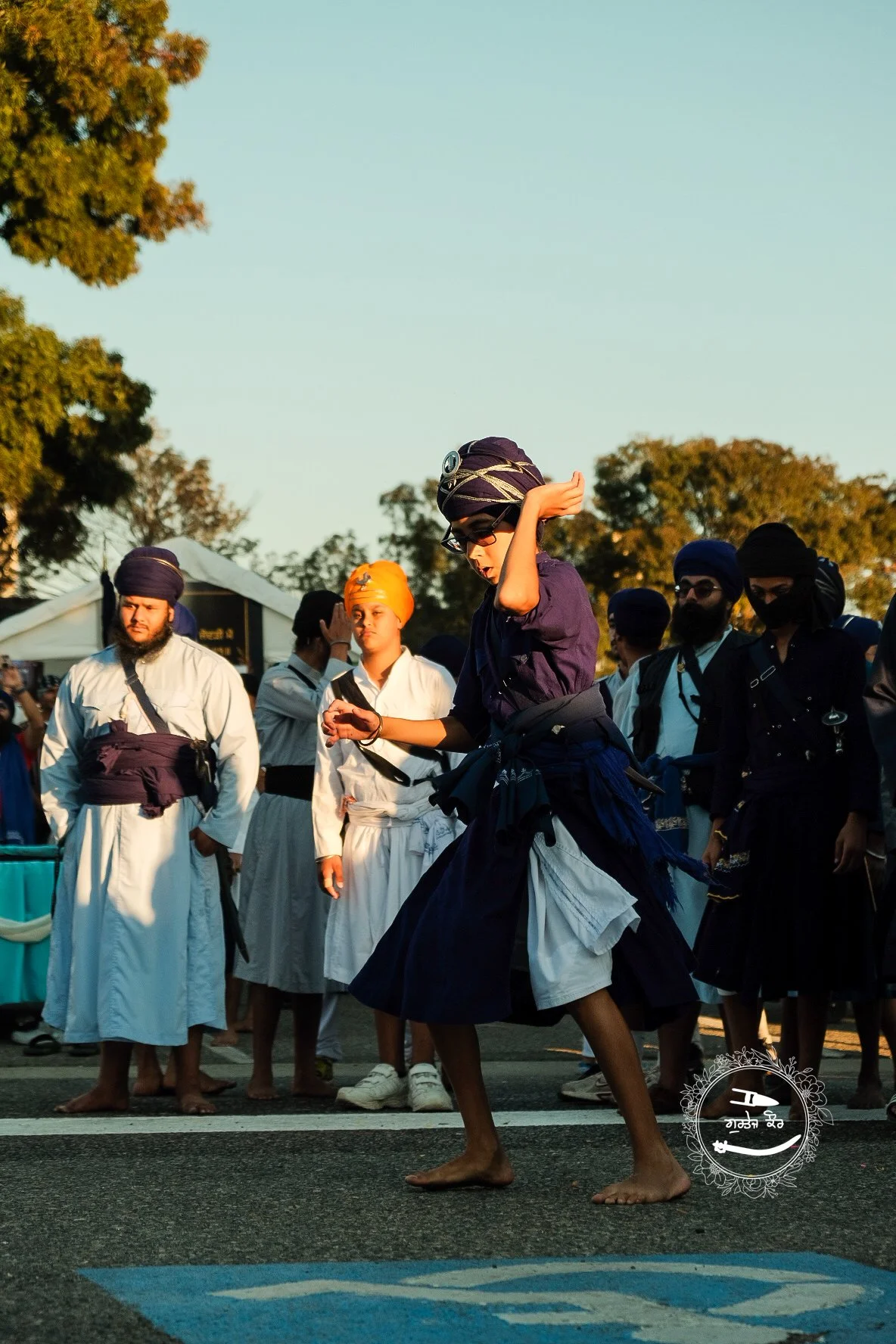 A young man dressed in traditional Sikh attire performs a dance on the street, surrounded by a group of men in similar attire, during a cultural event or parade.