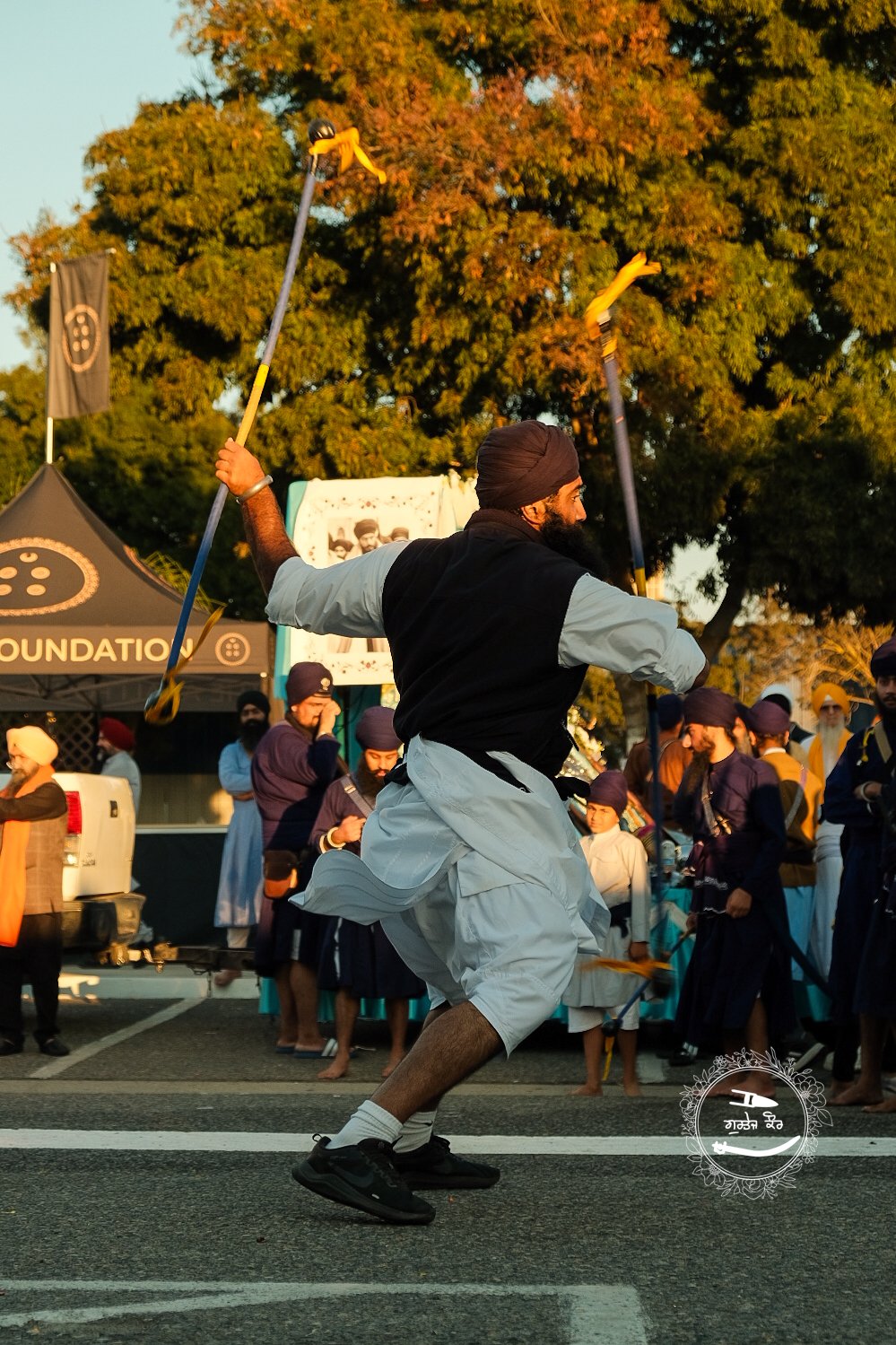 A man in traditional Punjabi attire performing Gatka martial art with two sticks during a cultural event, with a group of men in turbans and colorful clothing in the background.