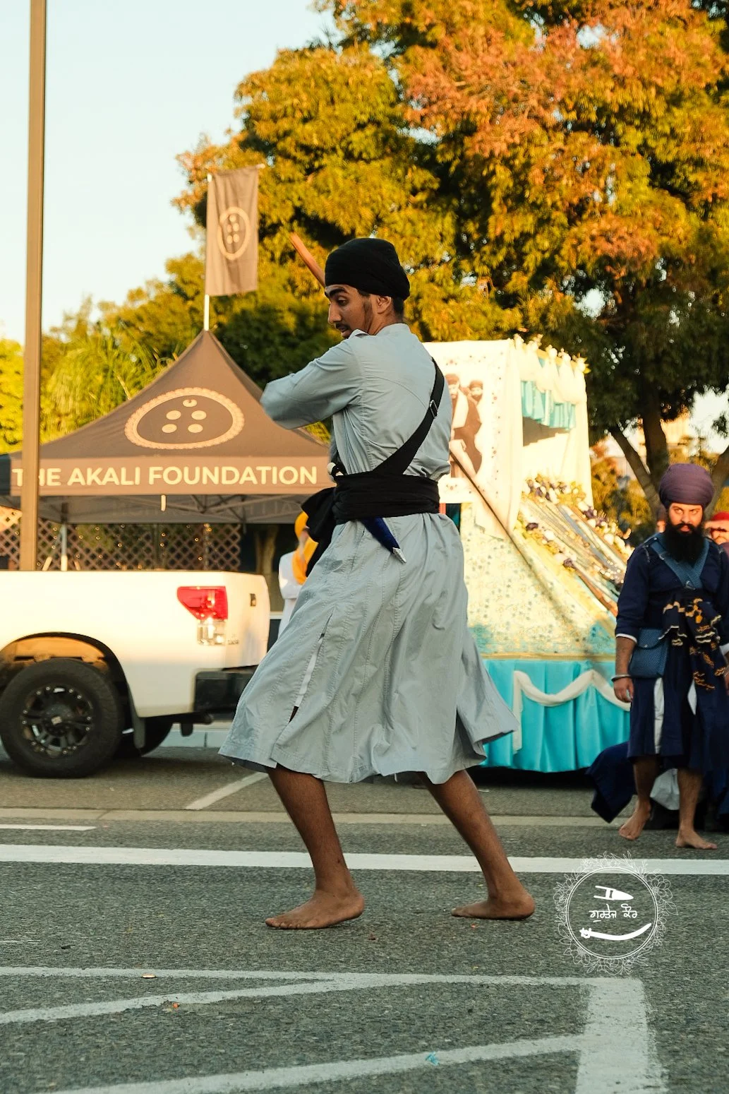 A man dressed in traditional Sikh attire and a black turban dancing barefoot on a street during a celebration or festival, with a tent, trucks, and other people in the background.