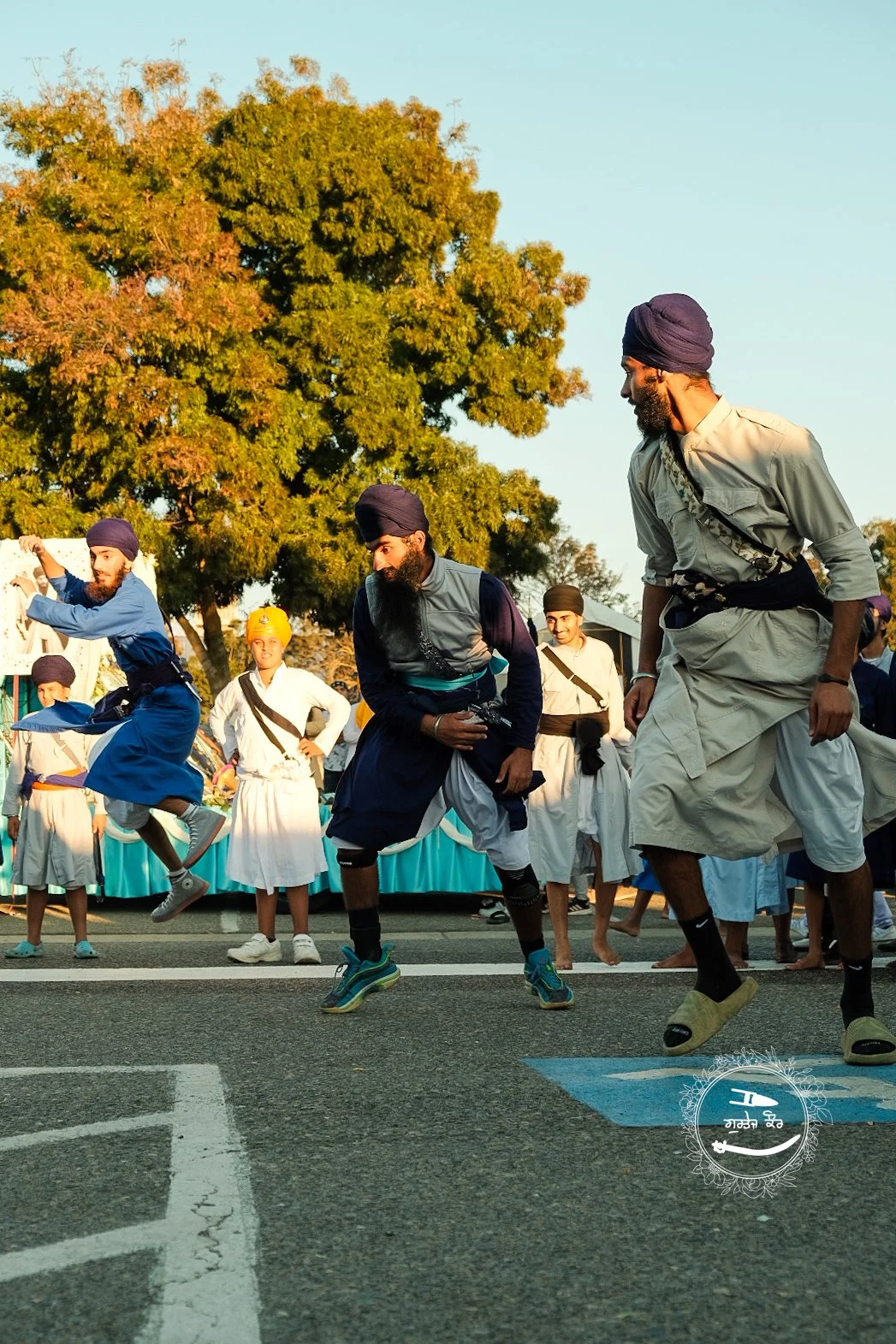 People dressed in traditional Sikh attire participating in a joyous dance performance on the street, with large trees and a clear sky in the background.
