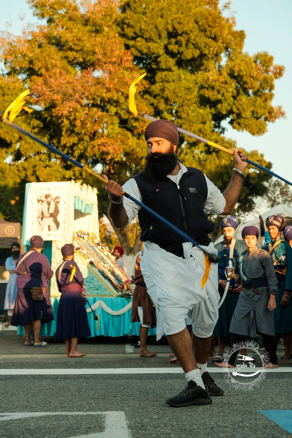 A man wearing a turban and traditional clothing, including a vest and white dhoti, performing a traditional dance with swords during a cultural parade. In the background, a group of people in traditional attire, and a decorated float with a framed ph