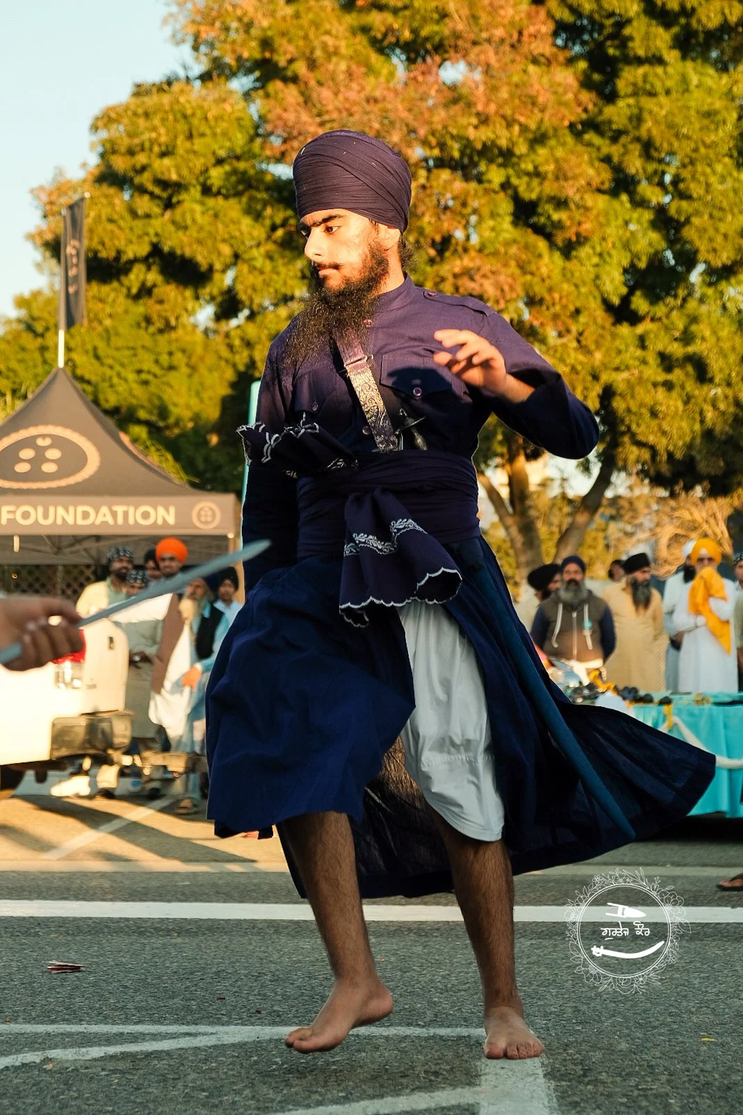 A man in traditional Indian attire, including a turban, performing a dance or martial art on a street during a festival, with onlookers and a tent in the background.