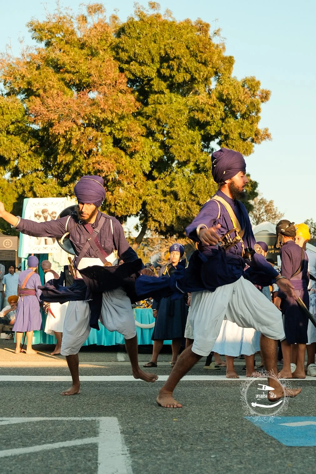 Two men dressed in traditional Sikh attire dancing barefoot on the street, with a large green tree and a crowd of people in the background.