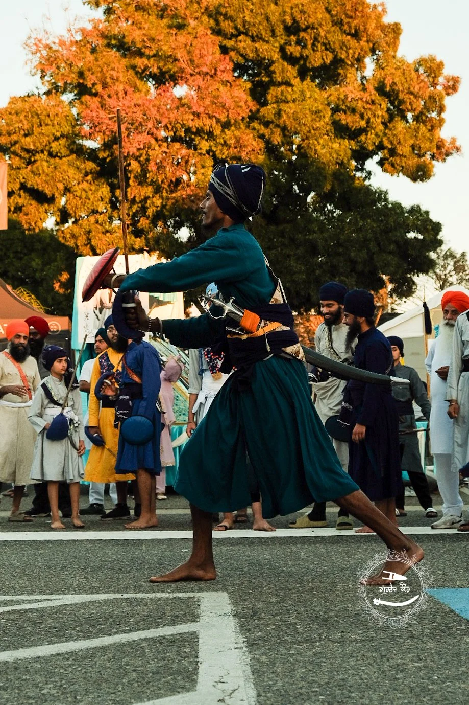 A man dressed in traditional Sikh attire performing a martial arts demonstration with a sword at a cultural event, with spectators in colorful clothing surrounding him and autumn trees in the background.