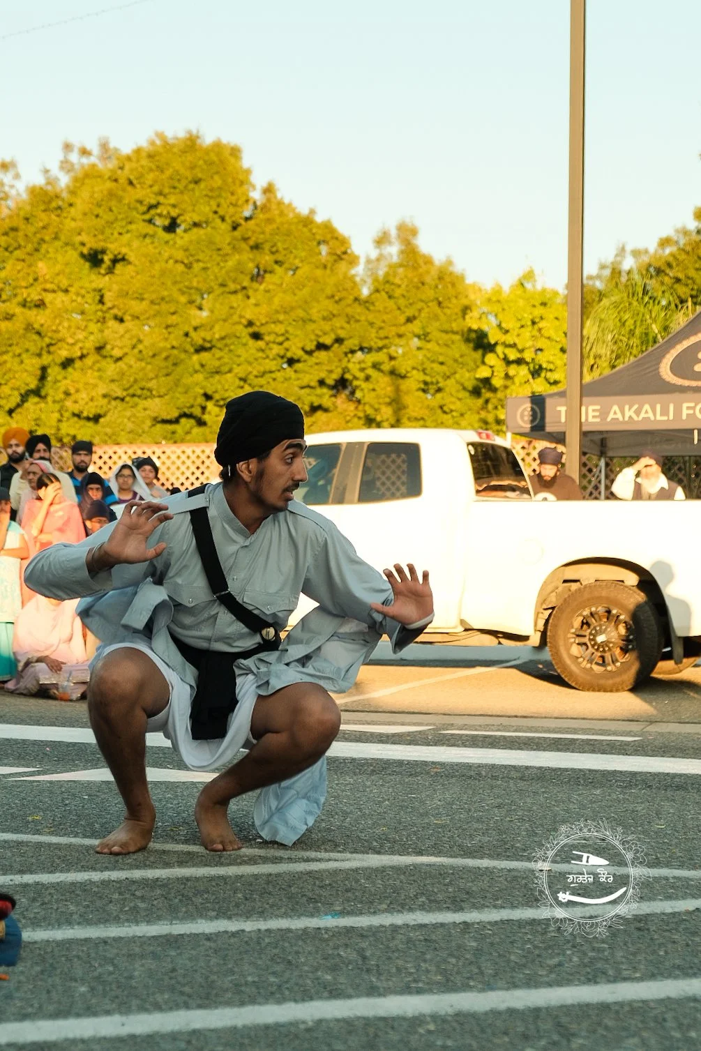 A man dressed in traditional attire crouches on the street, performing a martial arts pose with one hand extended and the other near his chest, in an outdoor setting with onlookers and trees in the background.