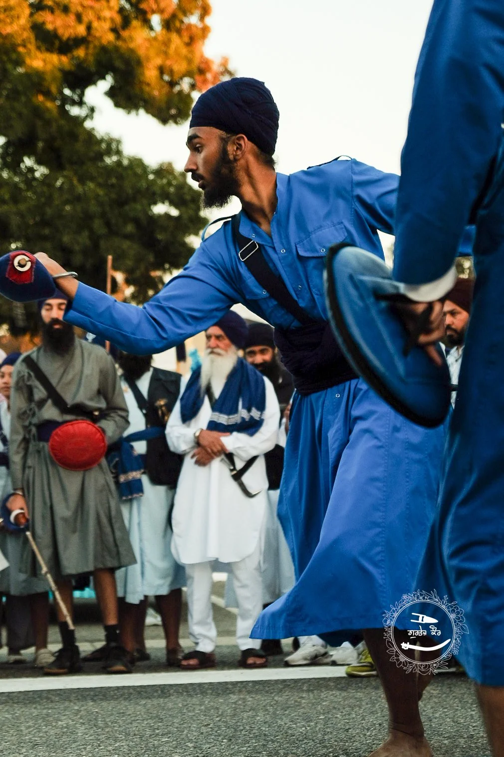 A man in a blue traditional Sikh attire, including a turban, performs a martial arts demonstration outdoors. Several onlookers, also in traditional Sikh clothing, observe him, with trees and a partly cloudy sky in the background.