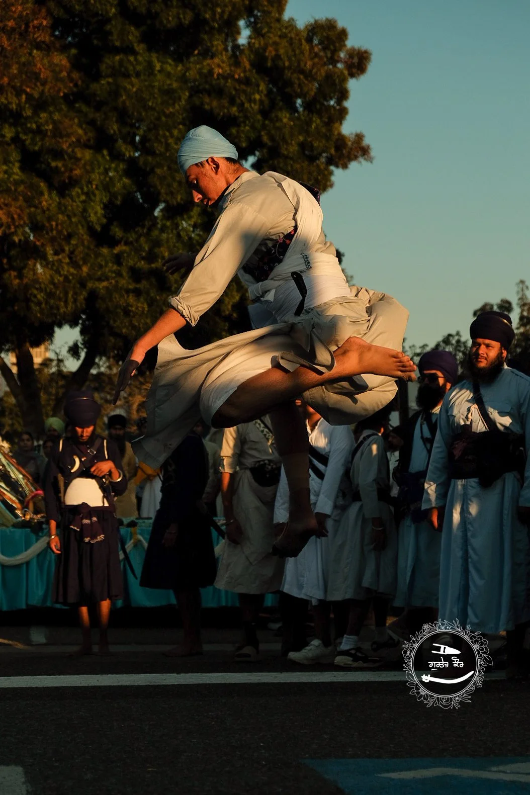A man dressed in white traditional attire jumping in the air during a cultural or religious festival, with a group of people in similar outfits standing behind him, some wearing turbans, in an outdoor setting during daylight.