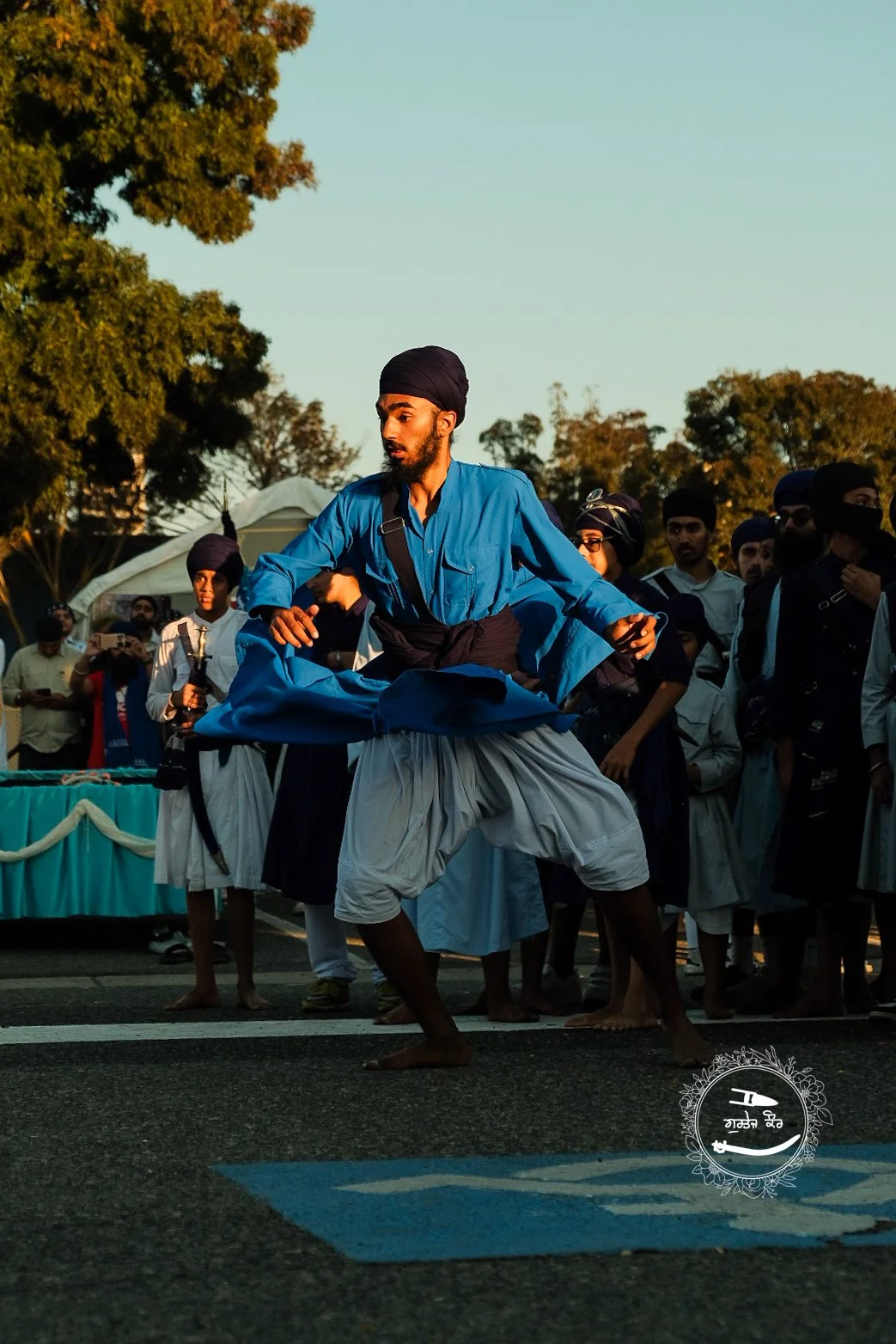 A man in traditional attire performing a dance or martial art in front of an audience during daylight, with trees and a canopy in the background.