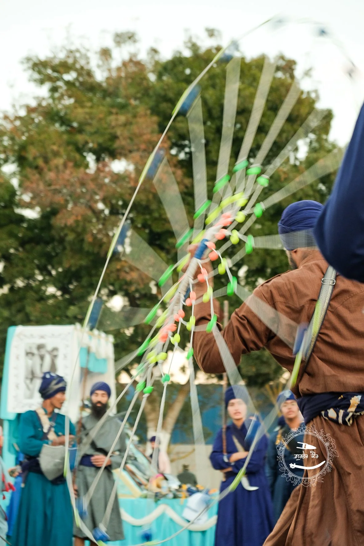 A man in traditional attire and a turban spinning a large, colorful kite at an outdoor event with several other people in the background and trees overhead.