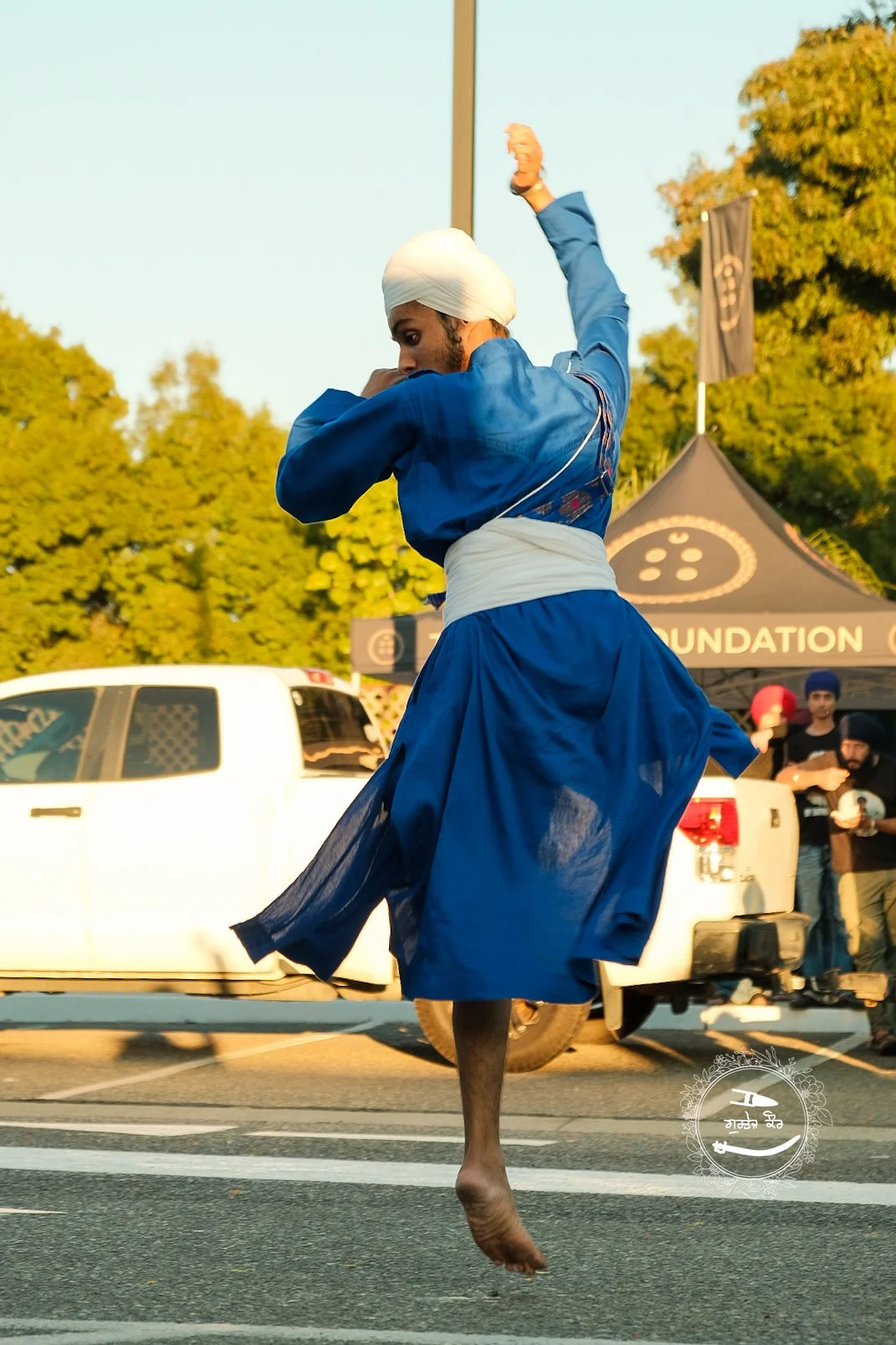 A man wearing a blue traditional outfit and a white turban is seen mid-air, jumping or dancing on a zebra-crossing street with a white vehicle in the background, during a public event with onlookers and a tent.