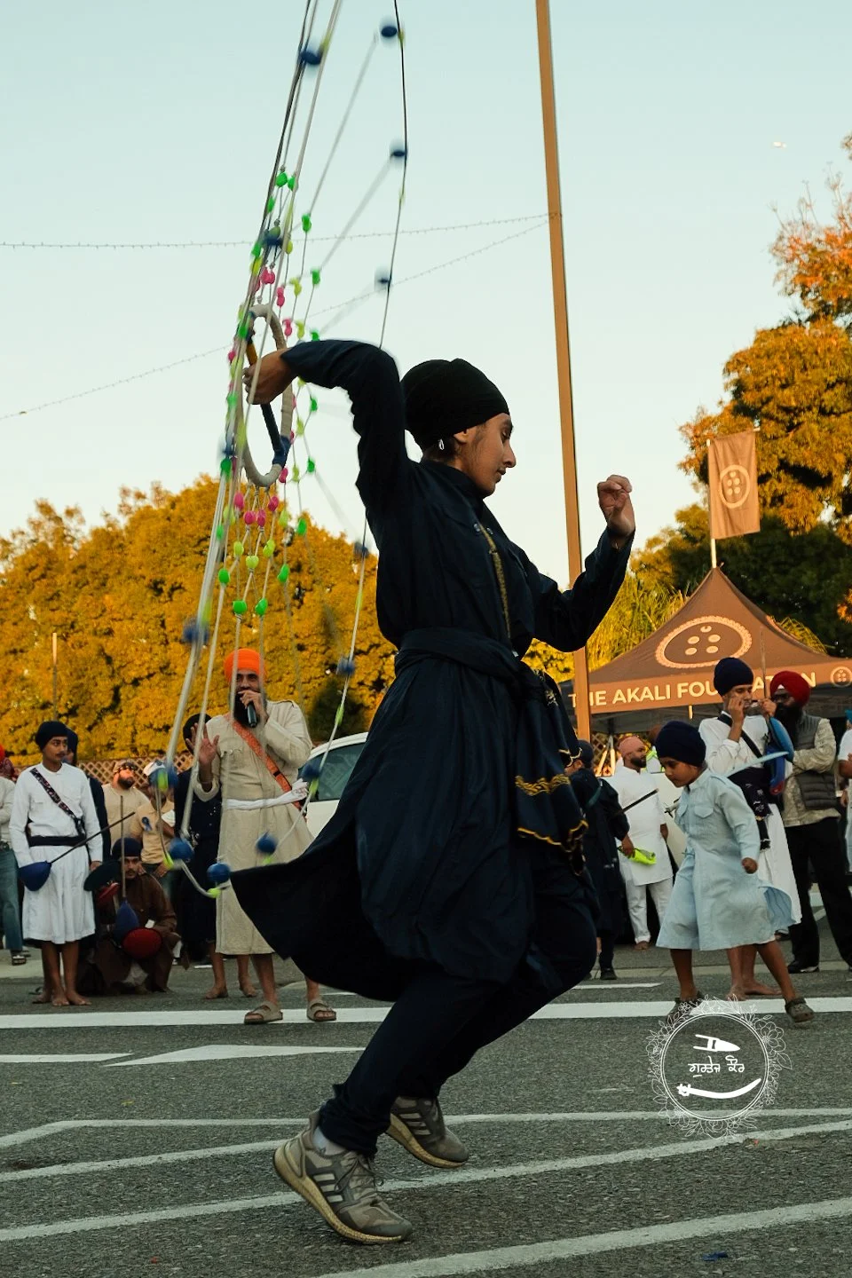 A man wearing a black turban and black attire is jumping while holding a large circular ring with a colorful rope and balls attached to it during a cultural event. Several people in traditional attire and children are gathered in the background, alon