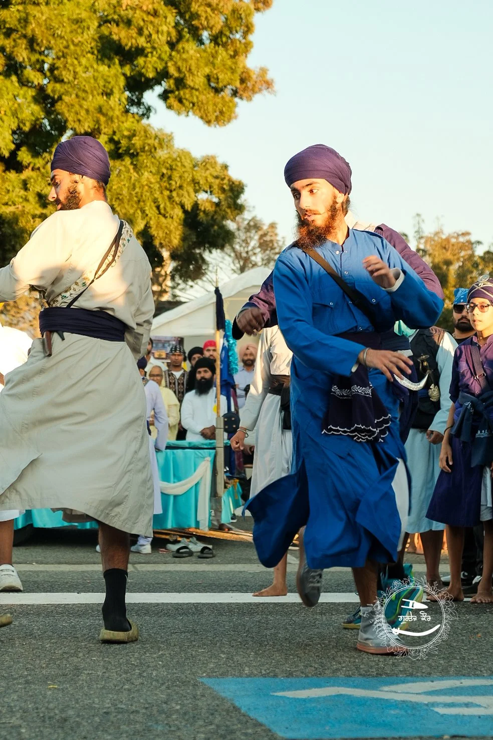Sikh men in traditional attire participating in a parade, wearing turbans and kurtas, with a crowd and trees in the background.