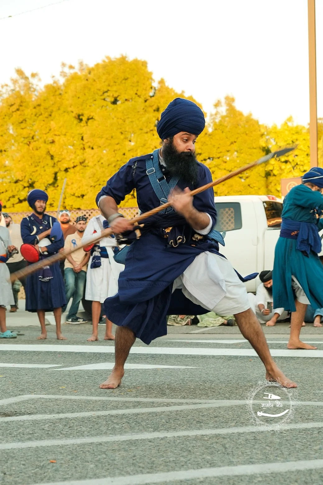 A man wearing traditional Sikh attire, including a blue turban and kurta, practicing Gatka martial art with a wooden stick on a street with yellow trees and other practitioners in the background.