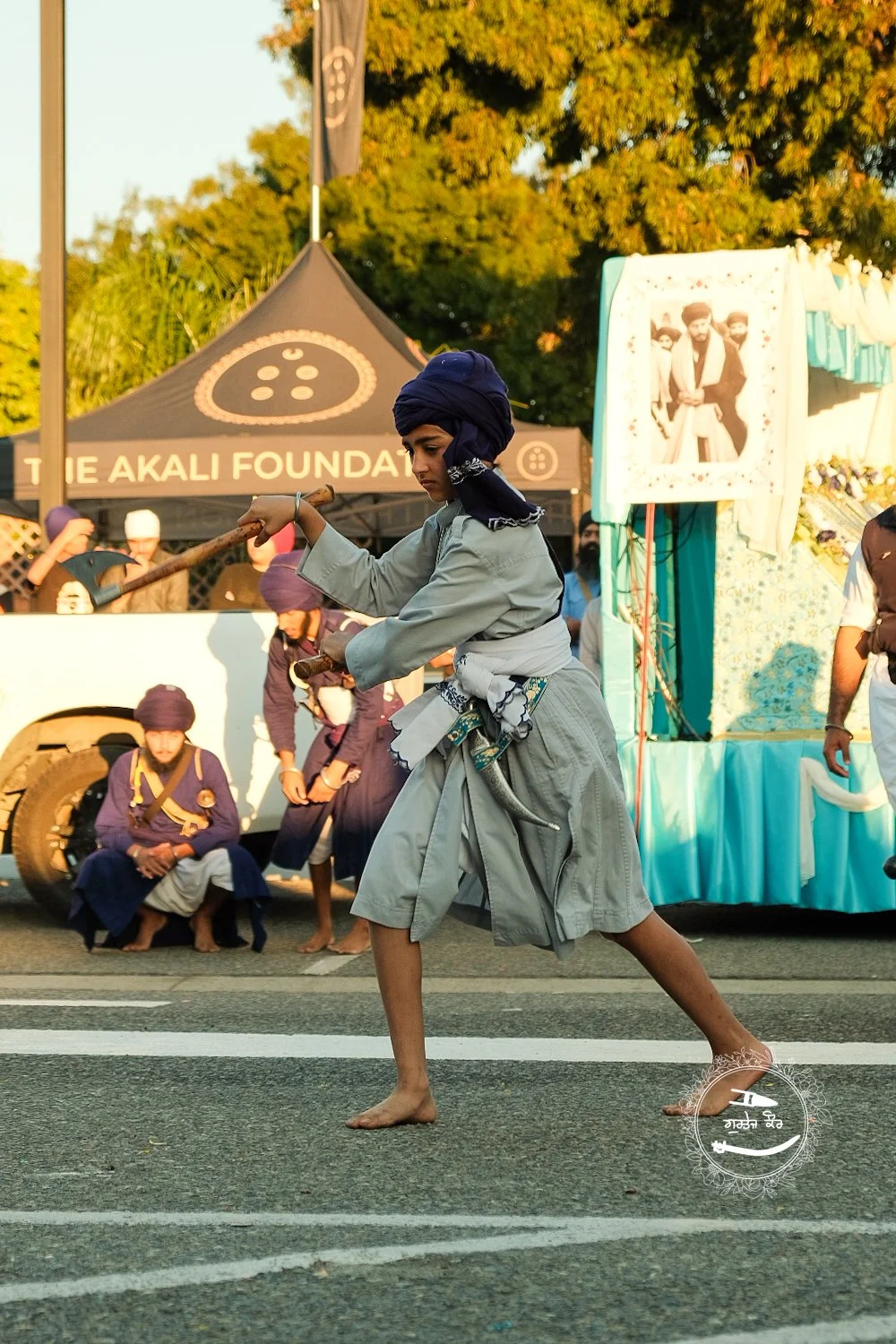 A young person dressed in traditional Sikh clothing performing a martial arts demonstration with a wooden stick on a street, with a parade float in the background during sunset.