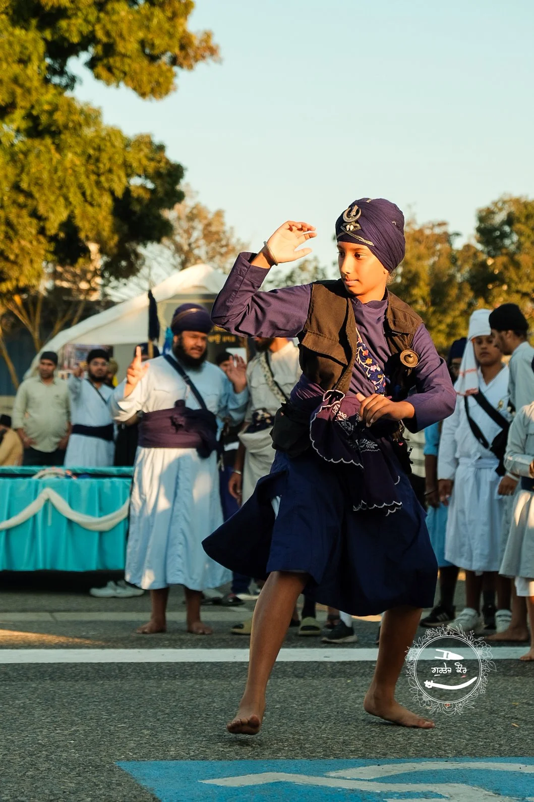 A young boy from the Sikh community, dressed in traditional attire with a turban and loose clothing, is performing a dance or martial art on the street as a crowd of men in traditional Sikh dress watch in the background during an outdoor event.