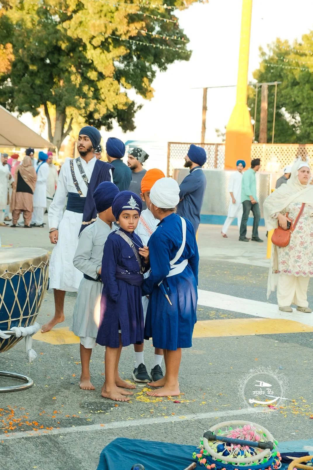 Group of Sikh boys and men dressed in traditional blue, gray, and white turbans and clothing, some barefoot, gathered and talking on the street during a festival or celebration, with others in the background and a drum and balloons visible.