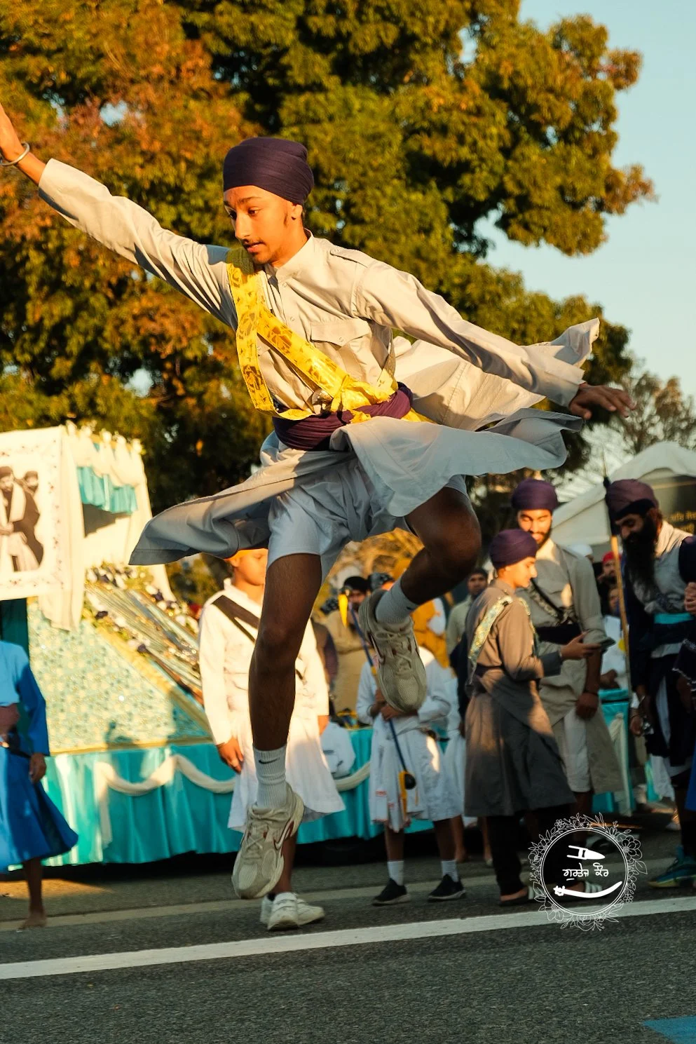 A young man in traditional Sikh attire, including a turban and kurta, is captured mid-air jumping during a festival or parade, with other participants and spectators in the background.