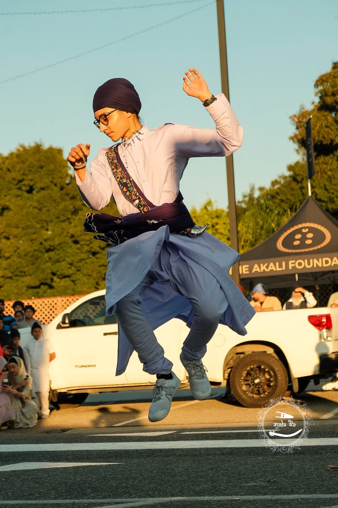 Person wearing a black turban, glasses, and traditional clothing jumping in the air during an outdoor event with onlookers and a black tent in the background.