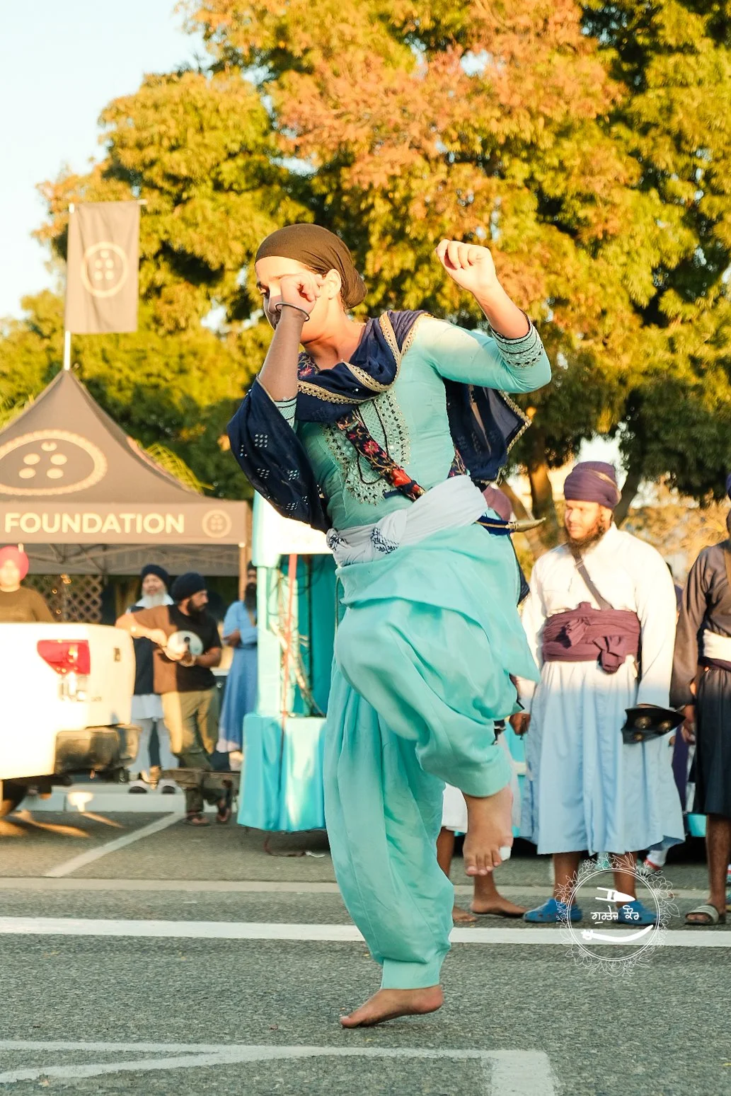 A person dressed in traditional Indian attire dancing barefoot on a street during a cultural event, with a large tree and a tent in the background.