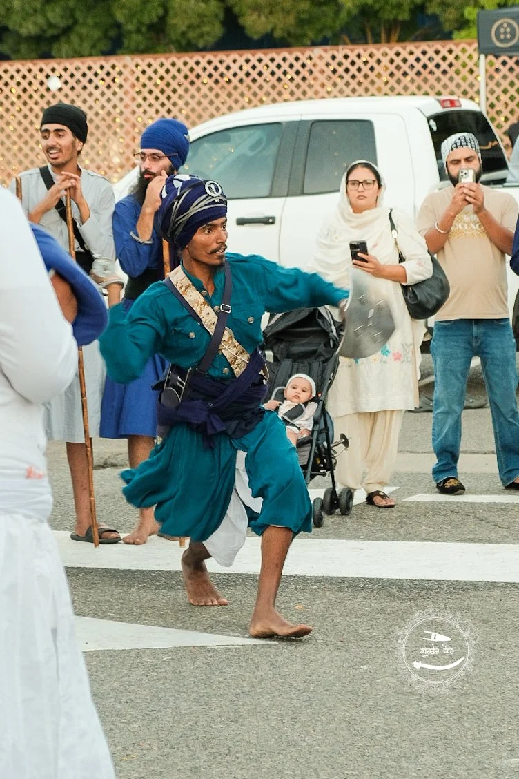A man dressed in traditional Sikh attire with a turban, performing a dance on a city street with spectators watching. Some people are taking photos or videos, and a baby in a stroller is among the onlookers.