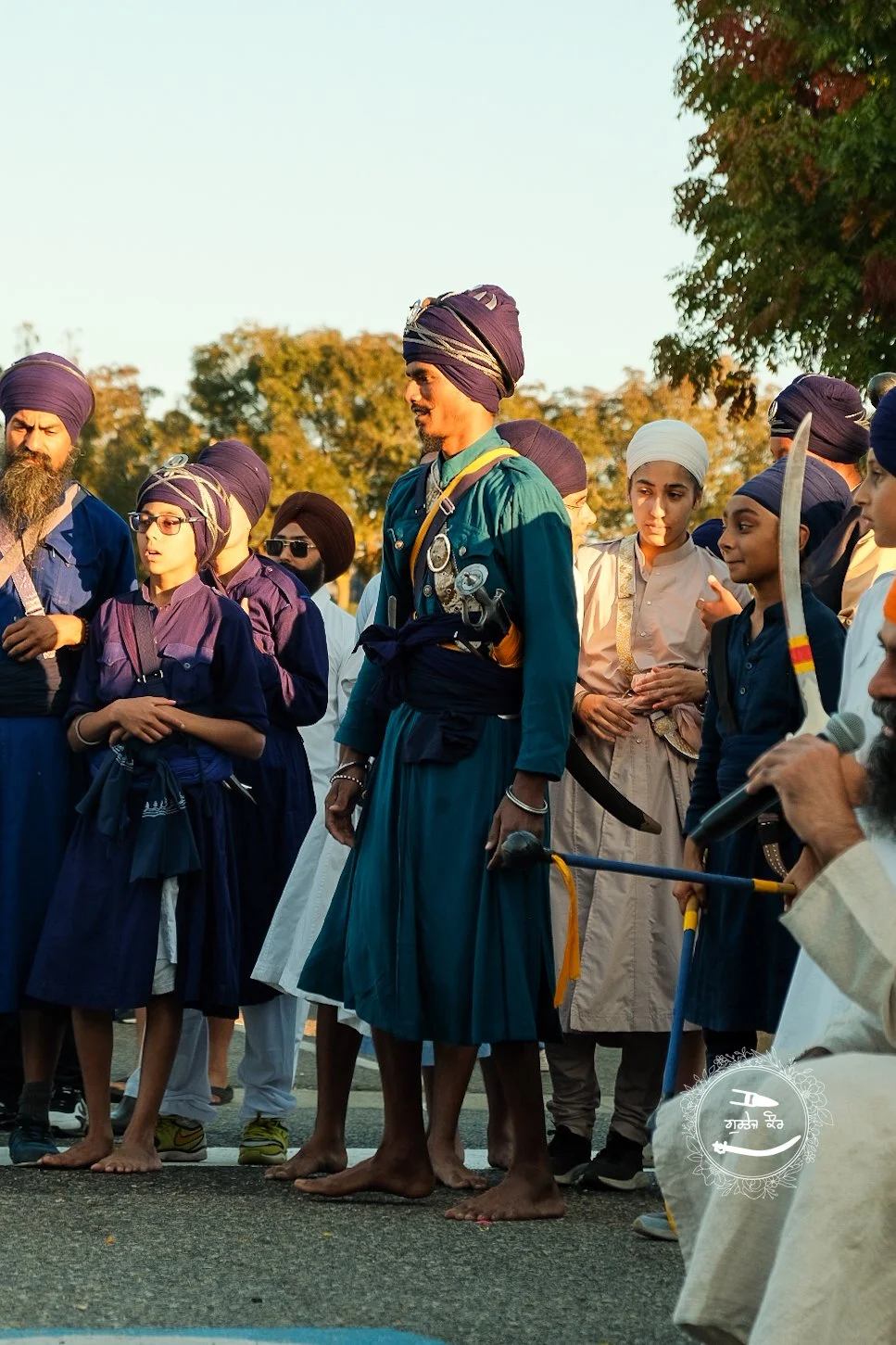 A group of Sikh men and boys gathered outdoors, some dressed in traditional attire with turbans and carrying swords, during a cultural or religious event in the evening sunlight.