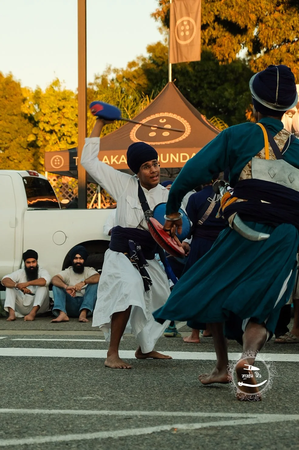 Two men dressed in traditional Sikh attire are performing Gidda dance on the street, while a group of seated men watch in the background during a cultural festival. A tent with the word 'Taka' is visible in the background.