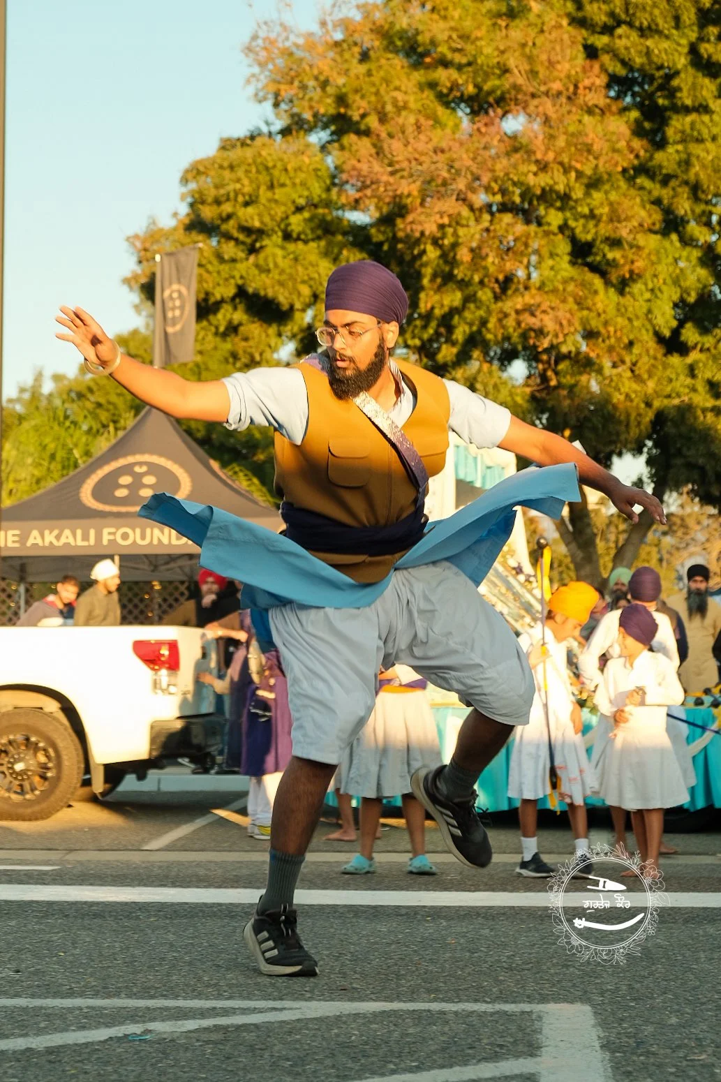 A man dressed in traditional Indian attire is dancing energetically on a street, with a group of children and adults in the background. The scene takes place during sunset, with trees and tents nearby.