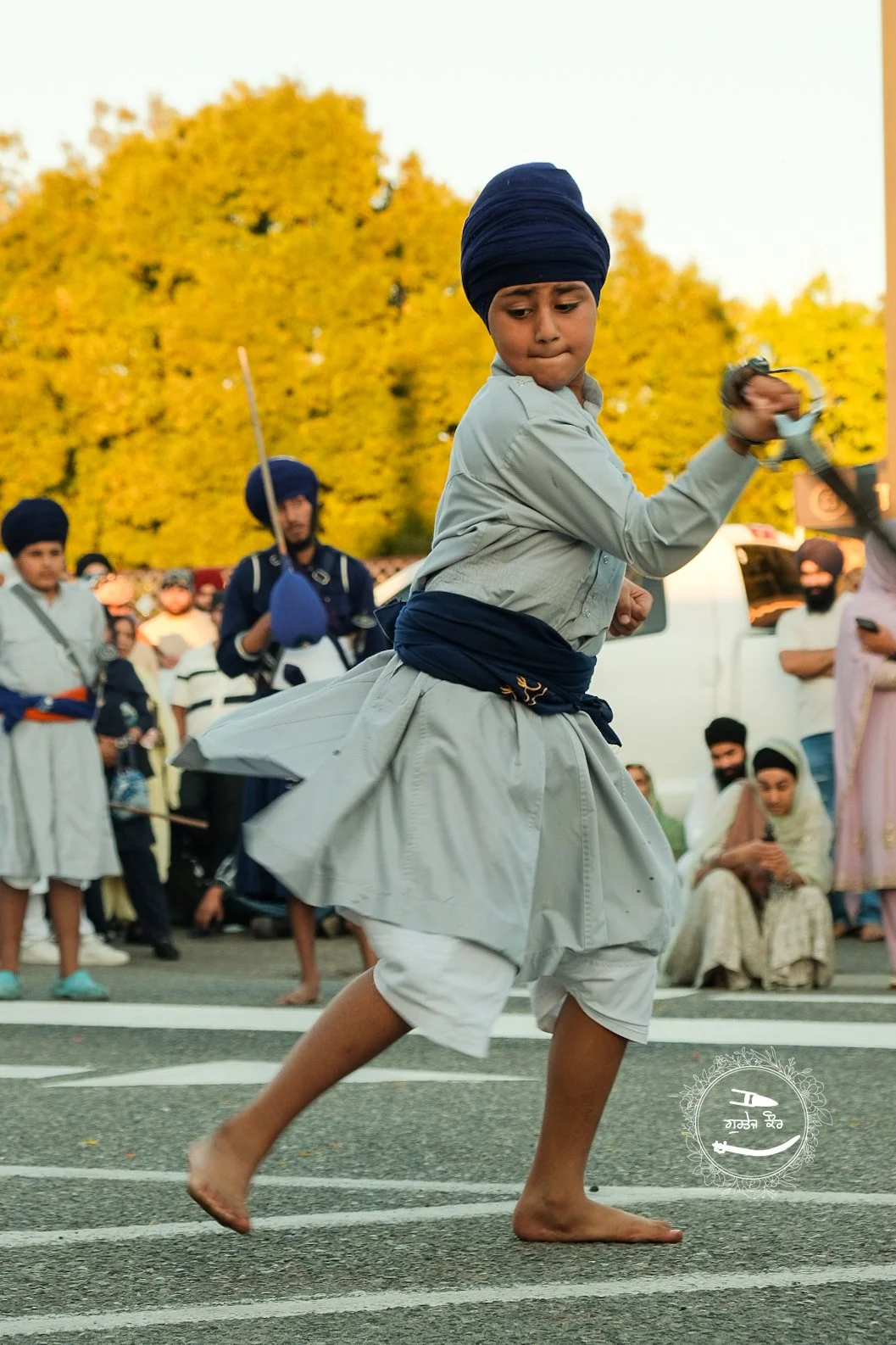 Young boy in traditional Indian attire performs a sword dance outdoors during a cultural festival, surrounded by onlookers.