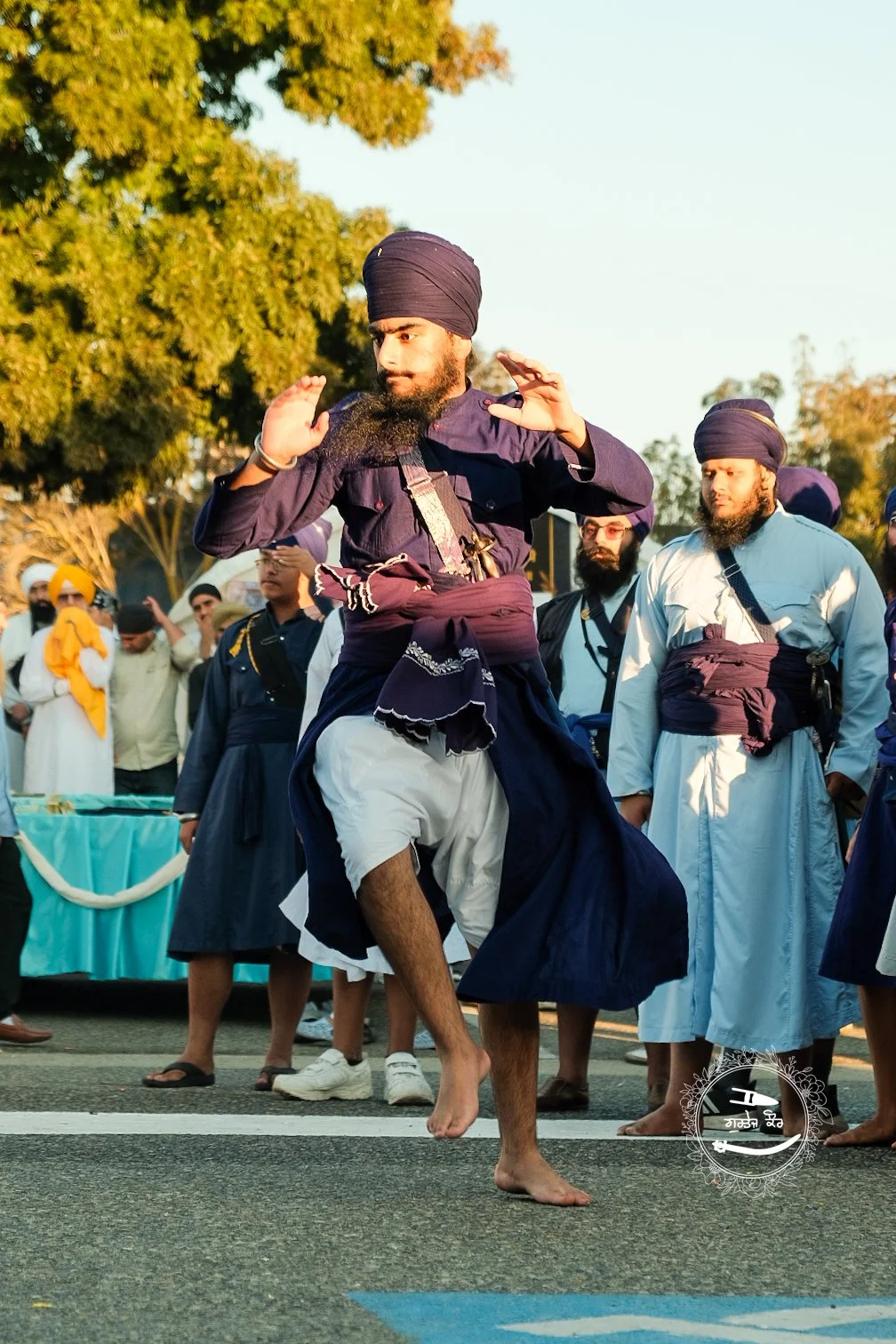 A man wearing traditional Sikh attire, including a turban and kurta, is dancing barefoot on the street during a cultural or religious celebration with a group of others dressed similarly in the background.