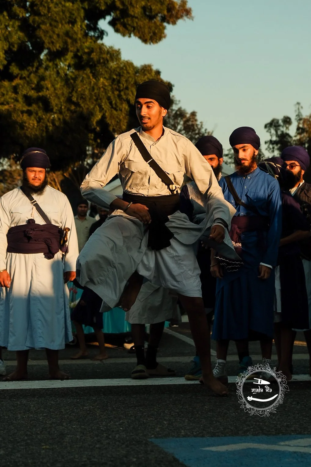A group of men in traditional Sikh attire participating in a martial arts demonstration, with one man performing a high kick while the others stand nearby, outdoors during the daytime.