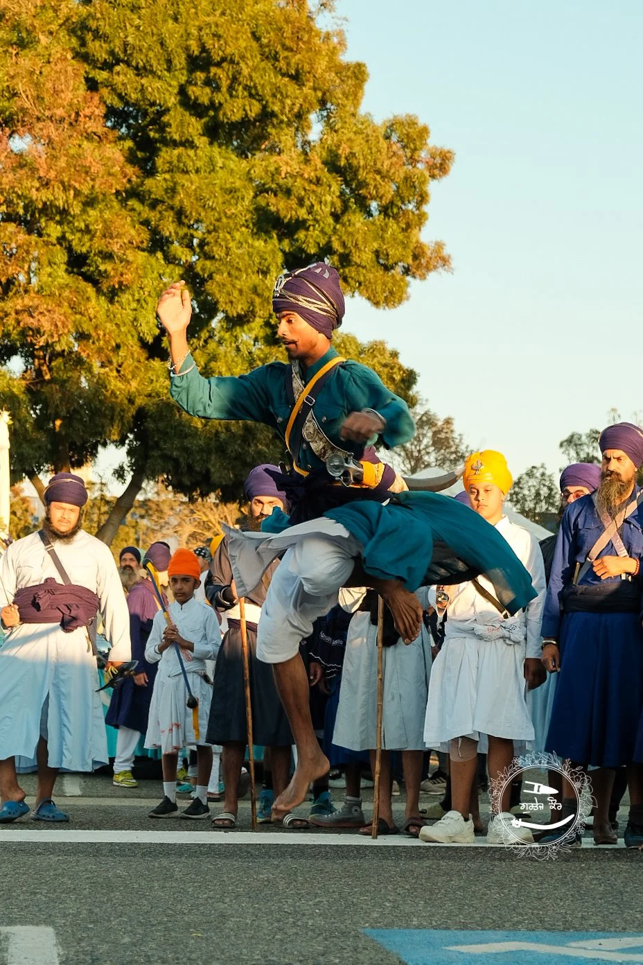 People participating in a traditional cultural event, with a man jumping with a sword while others look on, in a scene that appears to be a festival or ceremony in India.