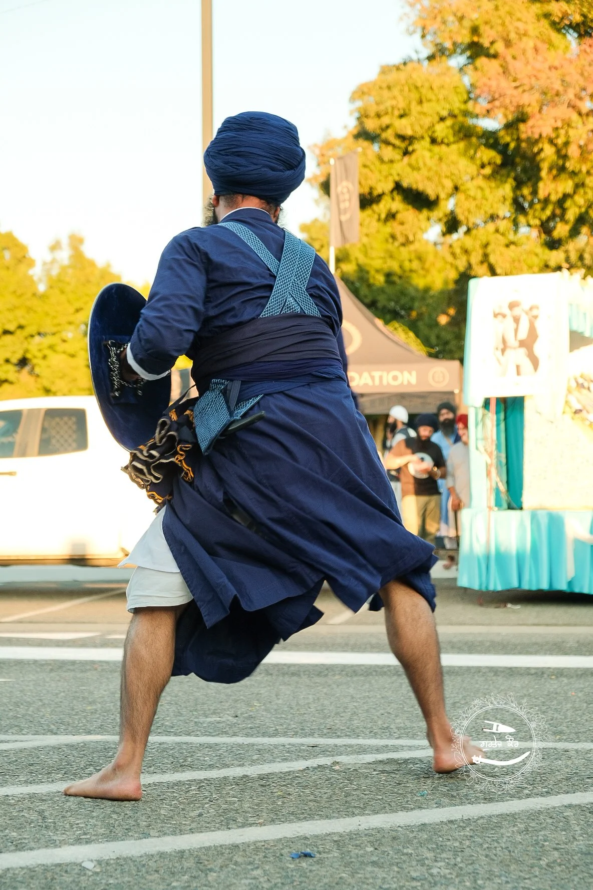 A man dressed in traditional Middle Eastern attire, including a blue turban and robe, holding a shield, performing a dance or martial arts move barefoot on a street at an outdoor event, with others and tents visible in the background.
