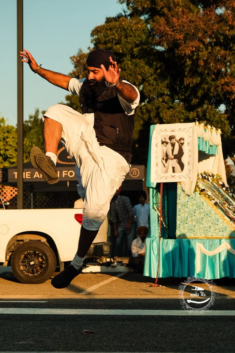A man wearing a turban jumping in the air with one leg bent and arms extended, in front of a float decorated with a photo of two men, during an outdoor event in the evening.