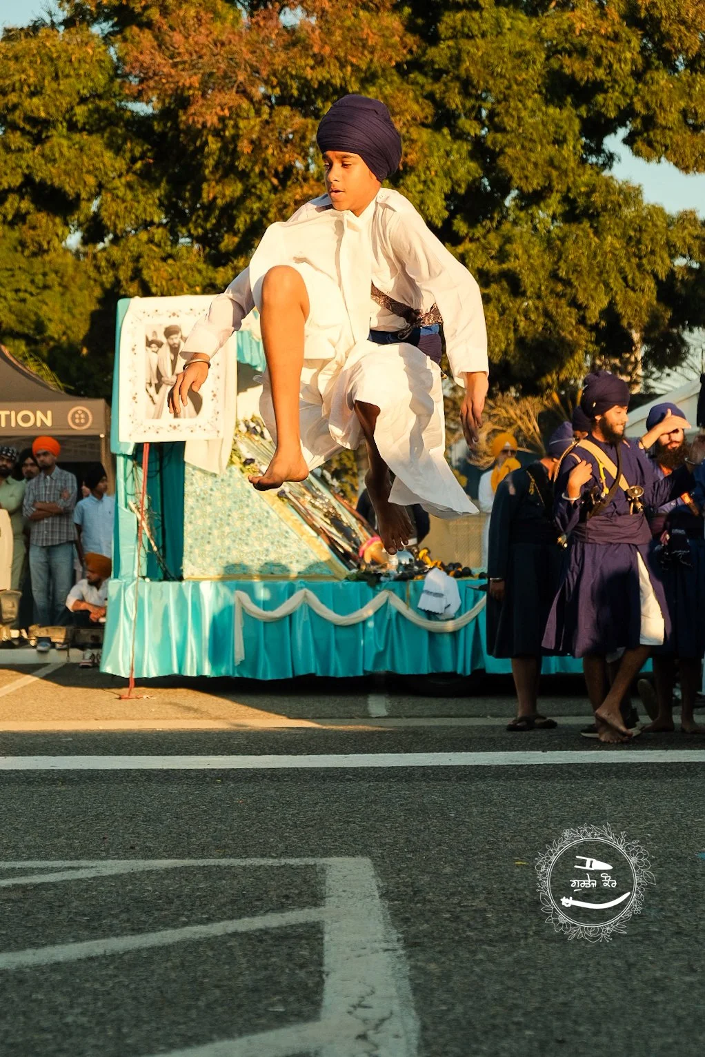 A young boy dressed in traditional white Indian attire and a black turban is mid-air, jumping or performing a stunt on a float during a parade. The float has a light blueish-green decoration with white drapes and features black-and-white photographs.