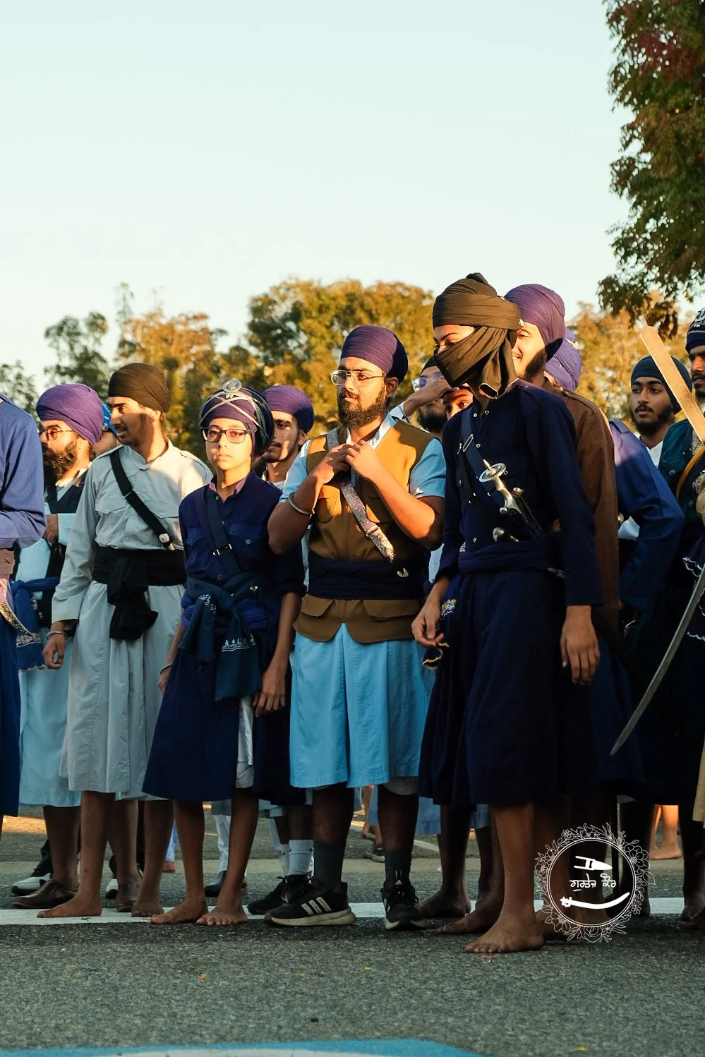 Group of Sikh men and boys standing outdoors, some barefoot, wearing traditional turbans and robes, during the golden hour sunlight.