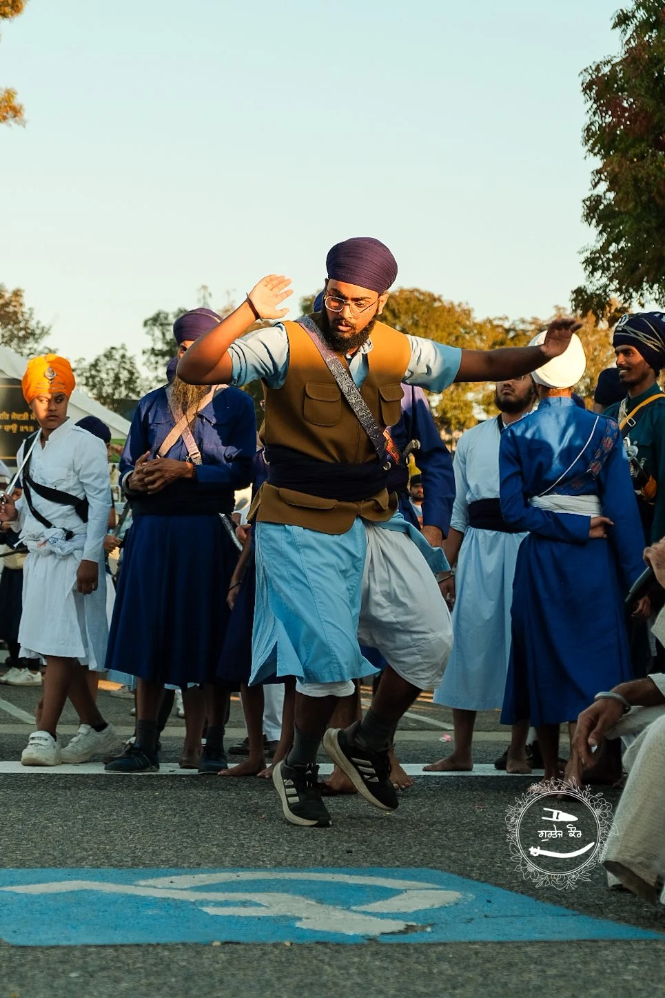 A man dancing with a bindi on his forehead, wearing glasses, a turban, and traditional Sikh attire, surrounded by other people also in traditional Sikh clothing and turbans, outdoors during daylight.