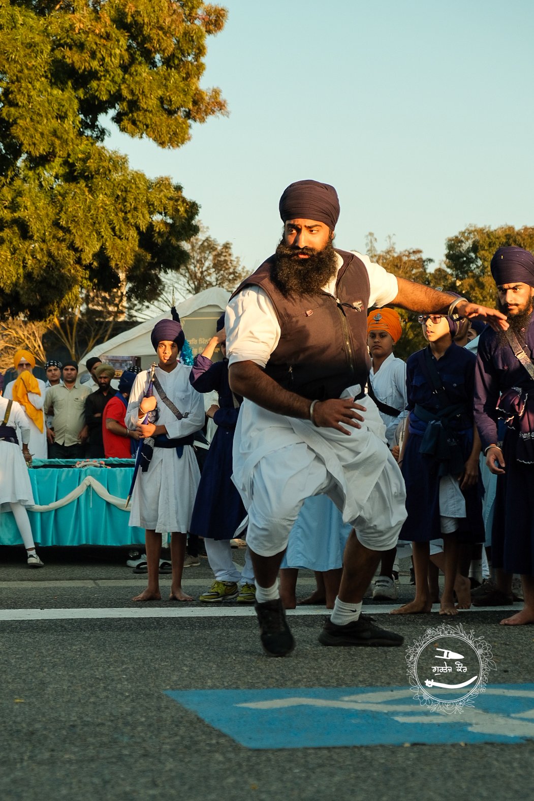 A man wearing traditional Sikh attire and turban dancing in a street parade, surrounded by other Sikhs and spectators with tents in the background.