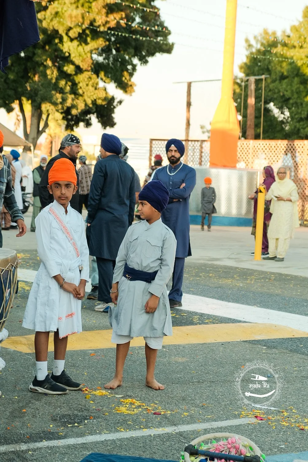 Two young boys dressed in traditional Sikh attire with turbans in orange and blue, standing on a street during a festival or religious celebration, surrounded by other people and festive decorations.