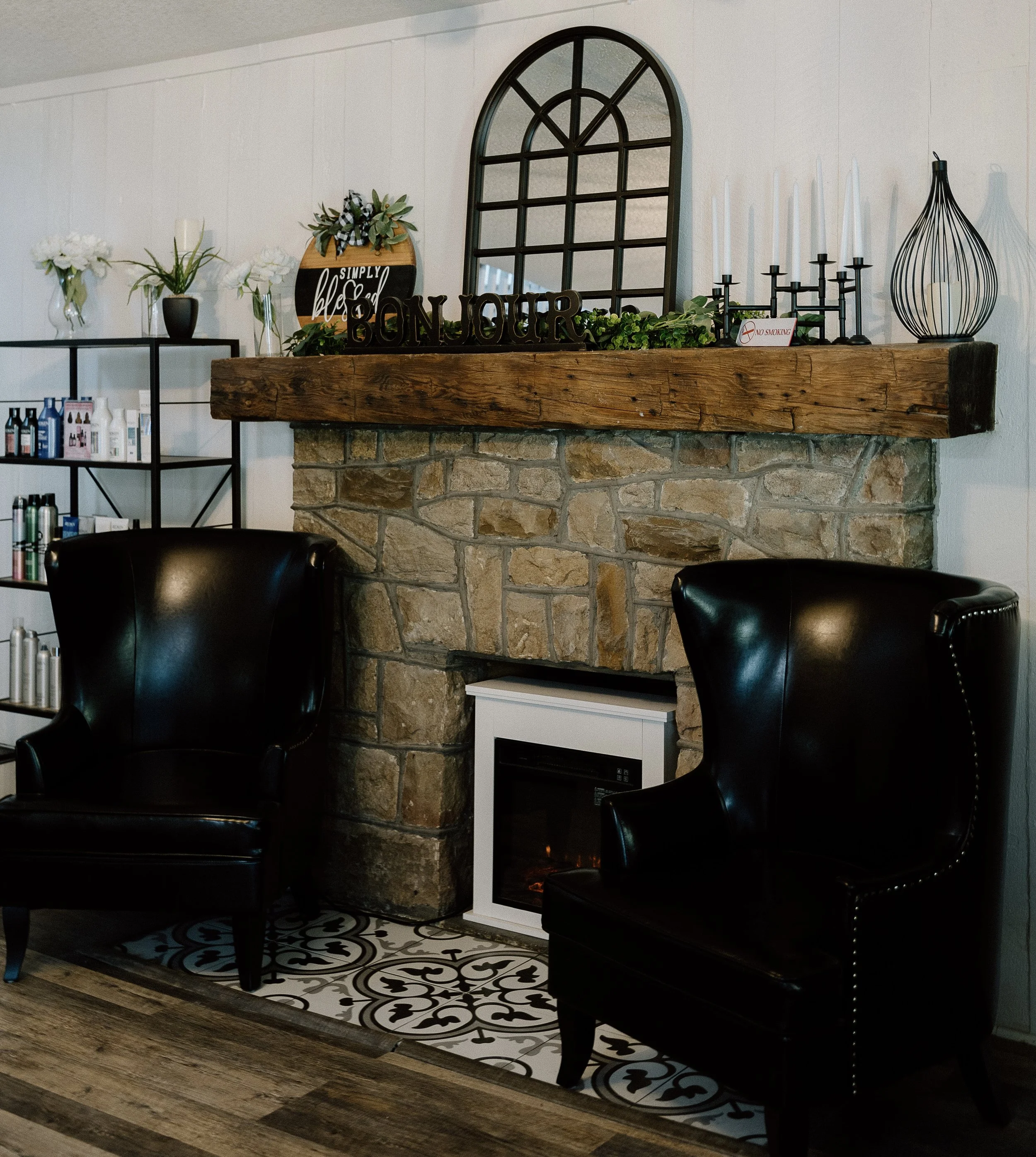 Living room fireplace with a wooden mantel decorated with green plants, a sign reading 'Simply Blessed', and black candle holders. Two black armchairs are positioned in front of the fireplace. To the left, a black shelf displays various bottles and products. The hearth is topped with patterned tiles, and the wall above the fireplace is decorated with a large surrounding mirror.