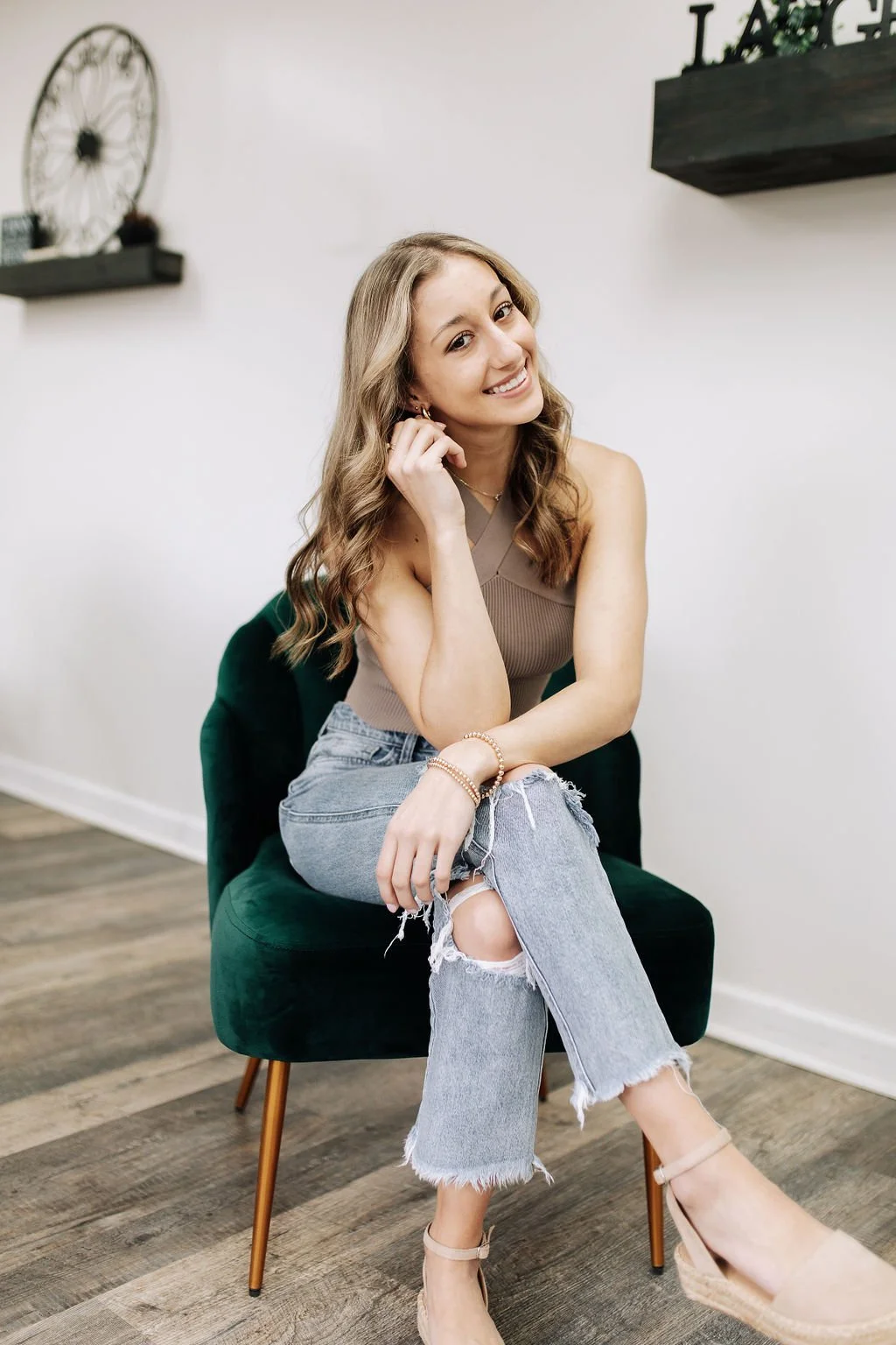 A young woman with wavy brown hair sitting on a green velvet chair, smiling at the camera, wearing a beige sleeveless top, ripped jeans, and nude heels, with a white wall and shelves in the background.