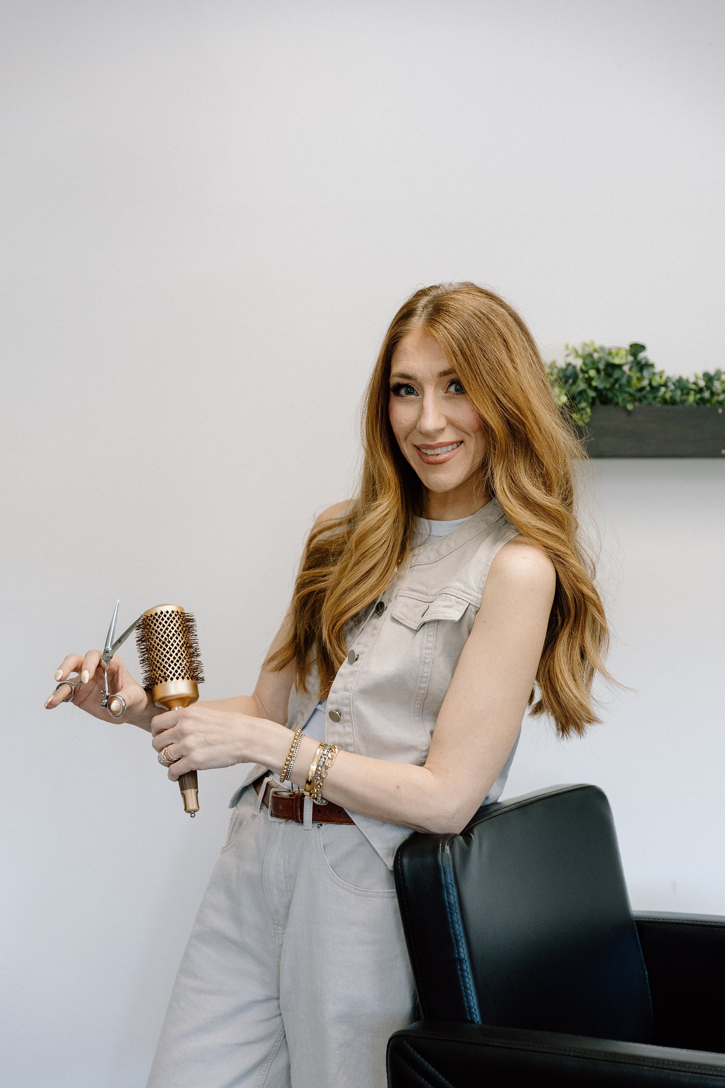 A woman with long red hair, wearing a beige sleeveless shirt and light-colored pants, sitting on a black chair with a high back, holding a hairbrush and scissors, in a minimalistic room with a white wall and a wall-mounted shelf with green plants.