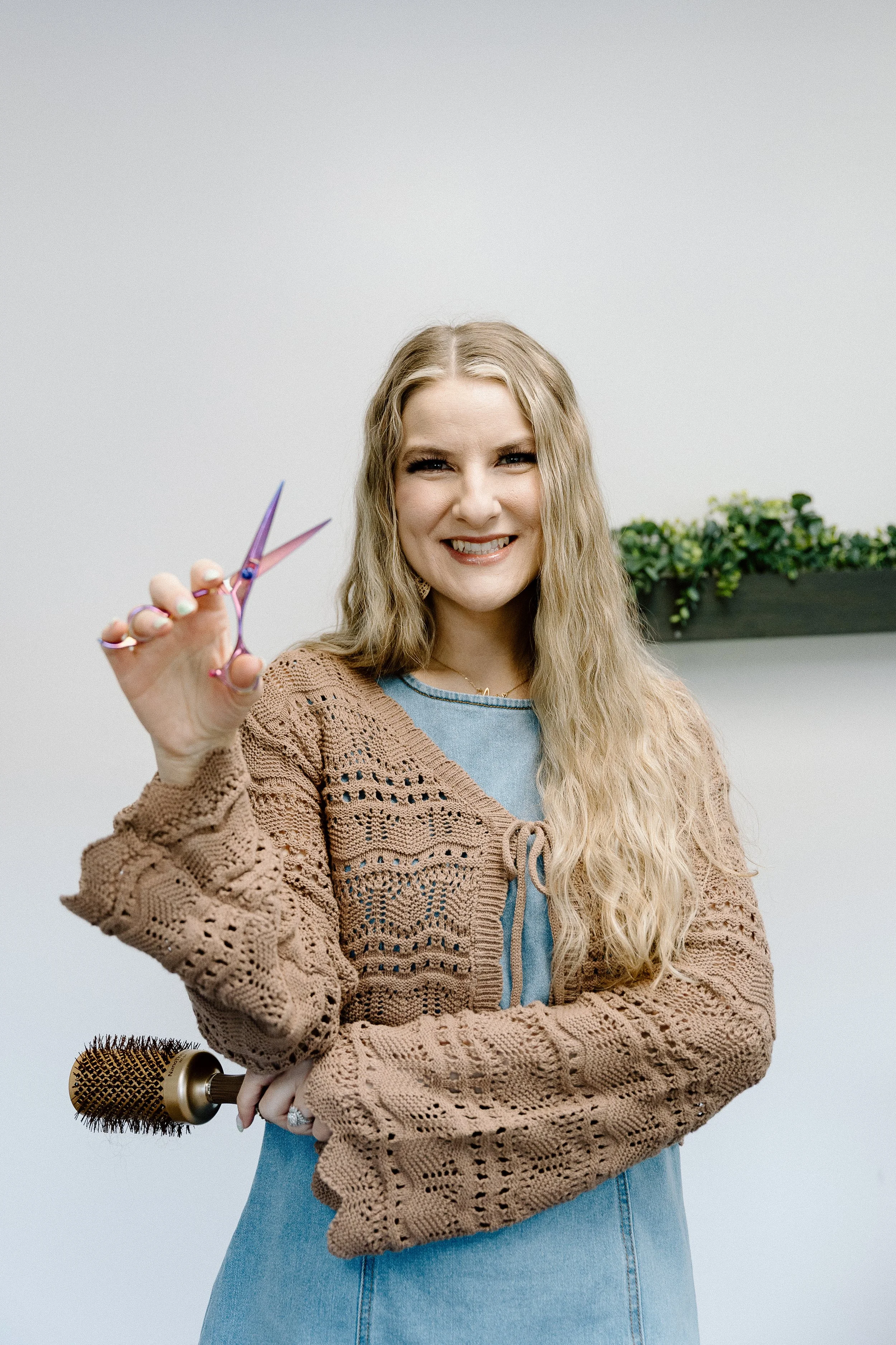A woman with long blonde hair holding a pair of pink scissors, standing in front of a plain light gray background with a potted plant and a hairbrush on a table.
