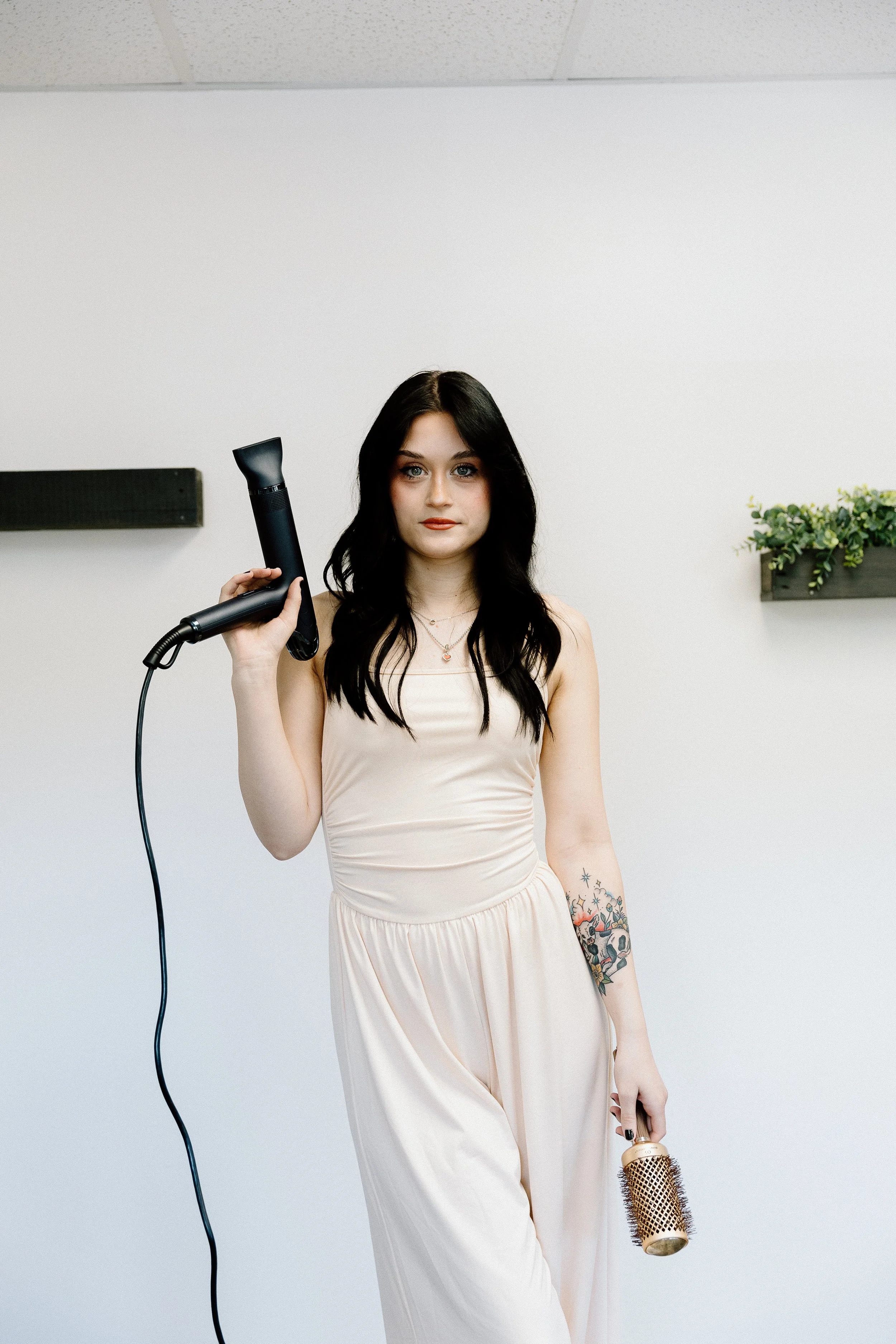 A woman with black hair, tattoos on her left arm, wearing a light-colored sleeveless dress, holding a hair dryer in her right hand and a hairbrush in her left hand, standing against a white wall with a black shelf and a small green plant.