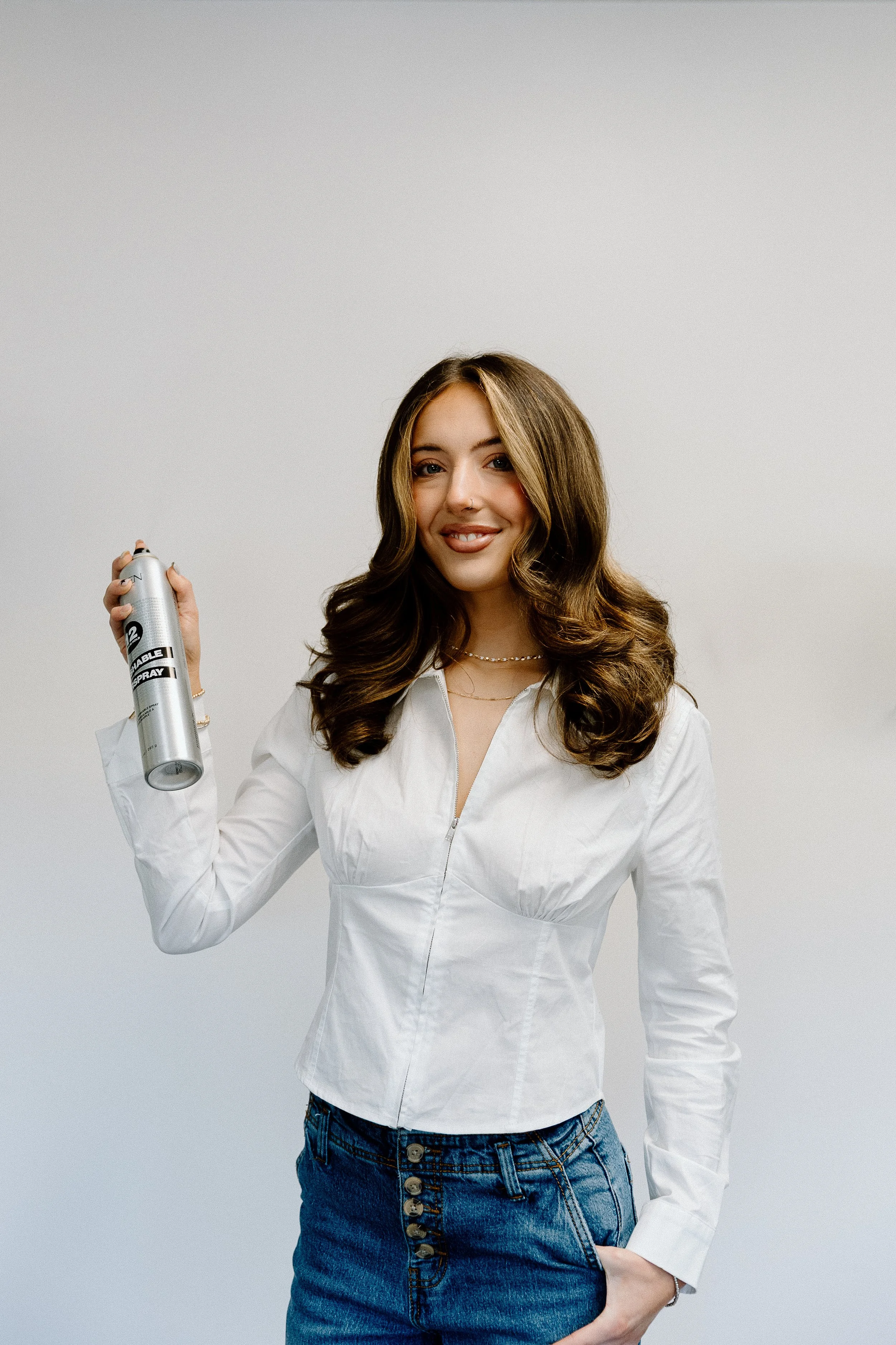 A woman with long, wavy brown hair and a light complexion holding an aerosol spray can in her right hand, wearing a white zipper jacket and jeans, standing against a plain light gray background.