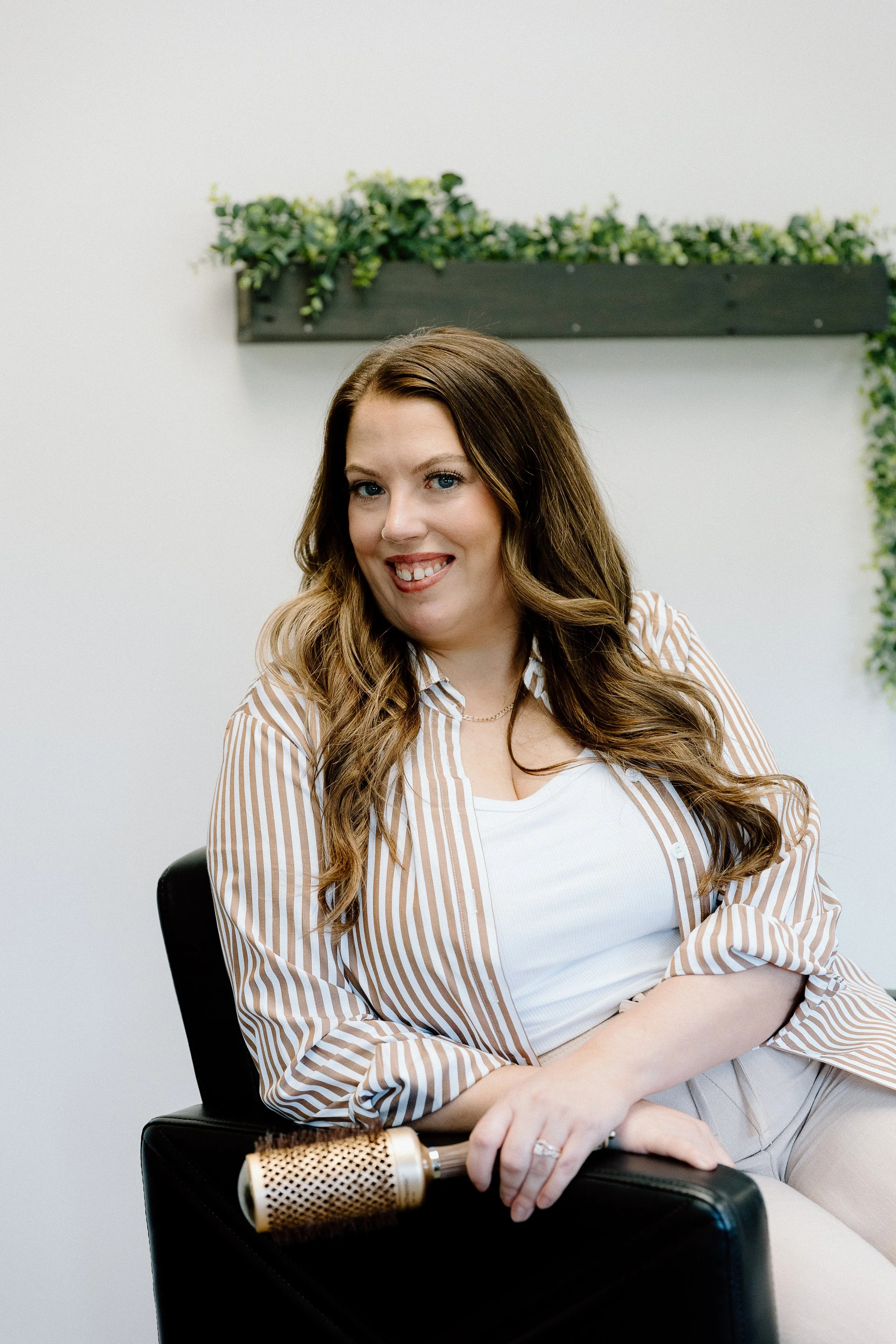 A woman with long brown hair and blue eyes, smiling, seated in a black chair, holding a gold microphone, wearing a white tank top and a beige and white striped shirt, in a room with white walls and a small green leafy plant on a shelf behind her.