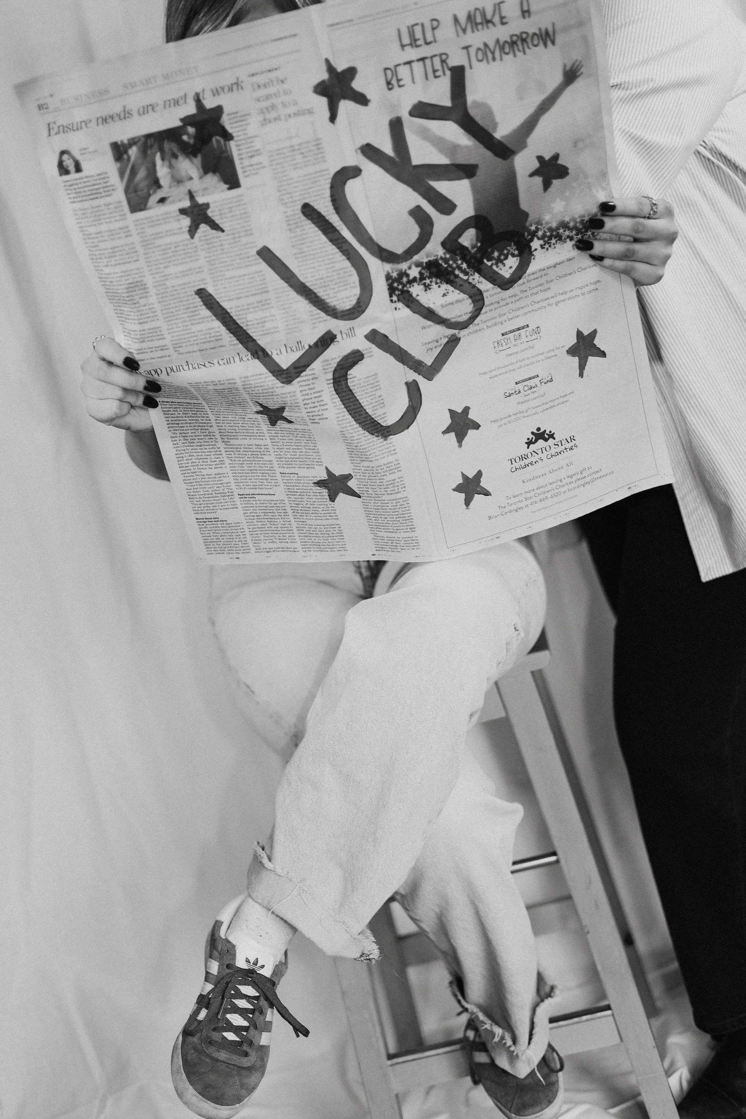 Person sitting on a chair, holding a newspaper with handwritten message 'Help make a better tomorrow' and colorful stars, with the word 'LUCKY' written in large letters.
